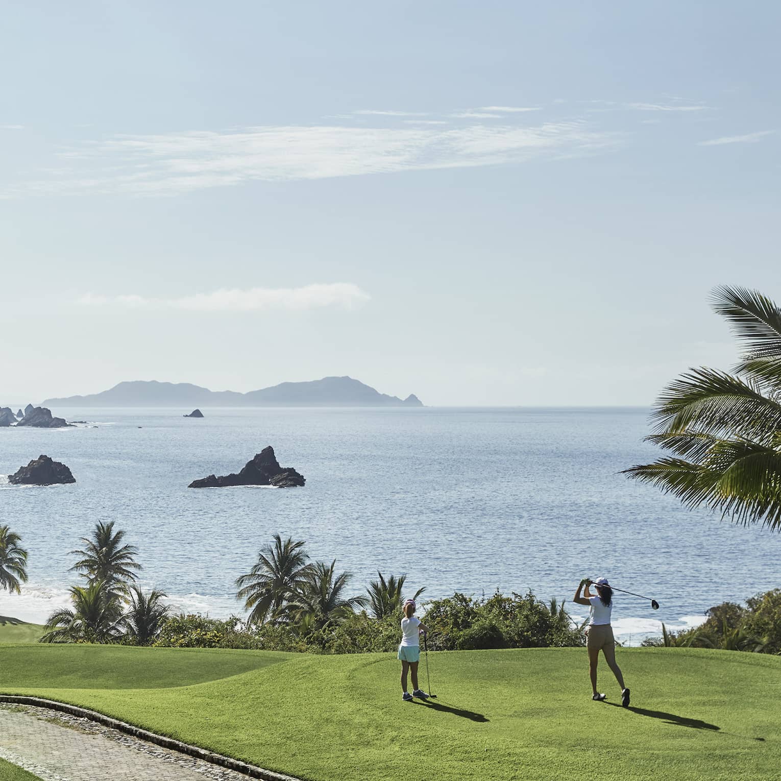 A guest swings from the tees on a hilly fairway bordered by palm trees and blue water with rocky outcrops as a child watches.