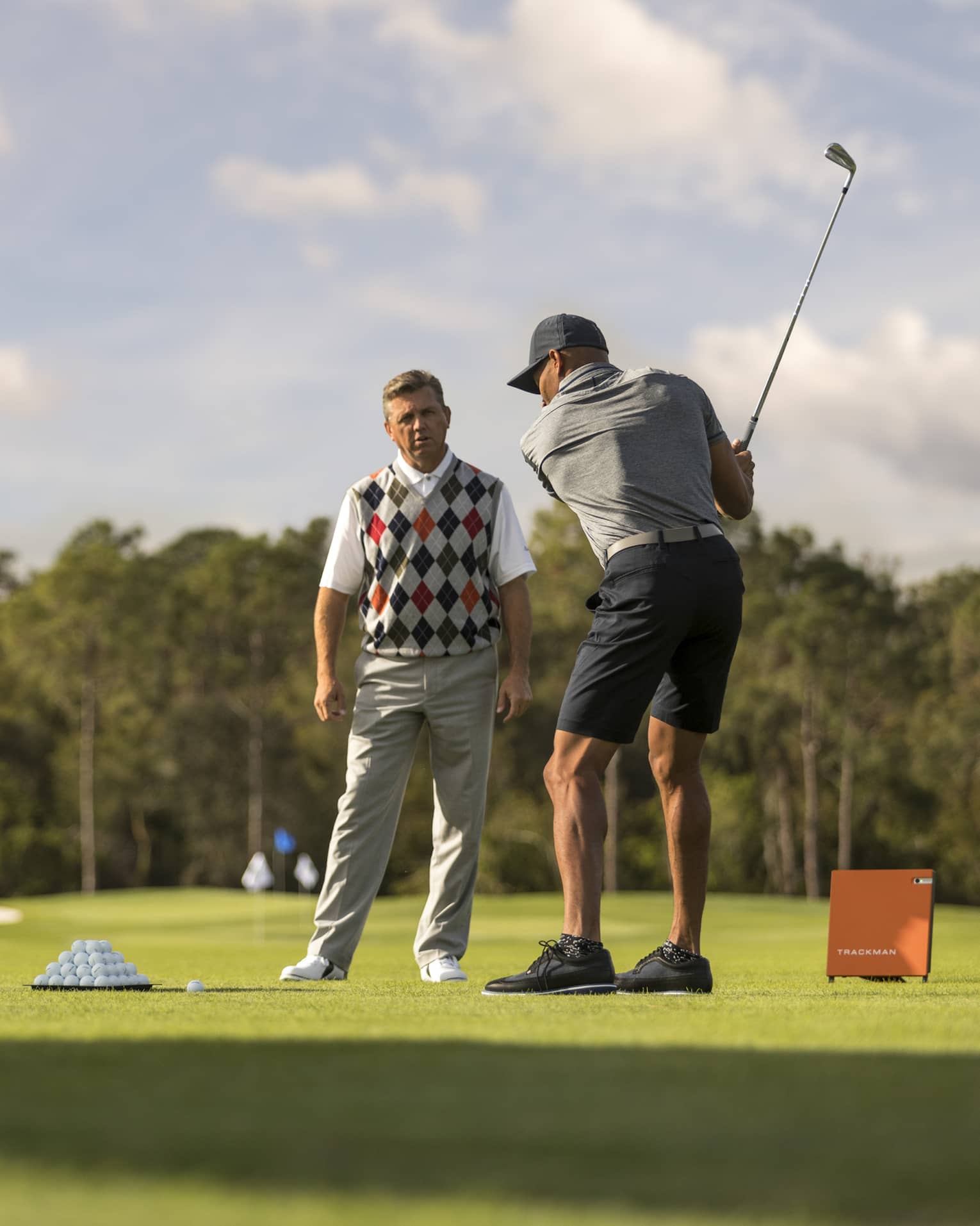 Against clear sky, one golfer looks on as another prepares to swing, a tray of balls and a stand of clubs nearby.