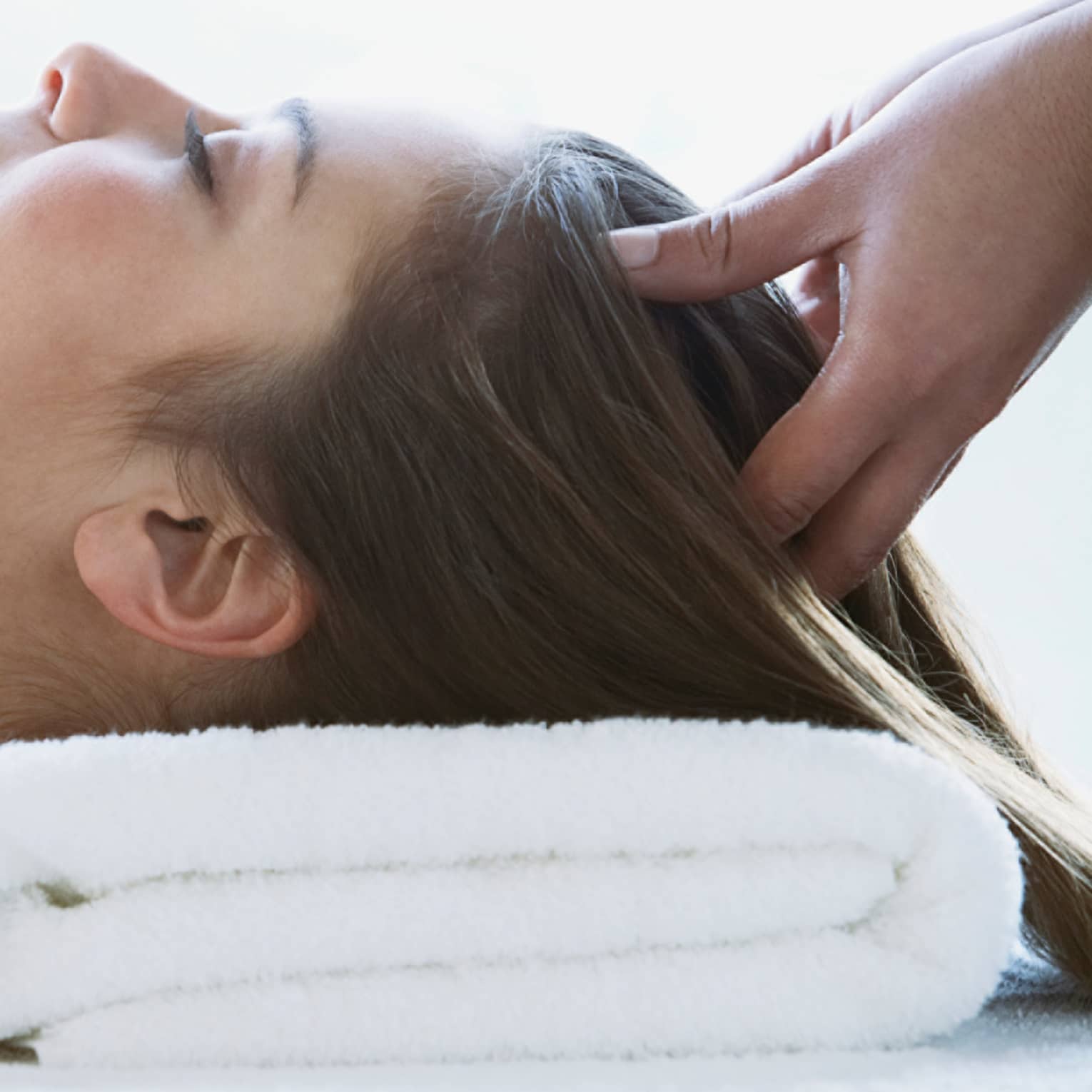 Woman lays on back with head on folded white towel, closes eyes as hands massage her scalp