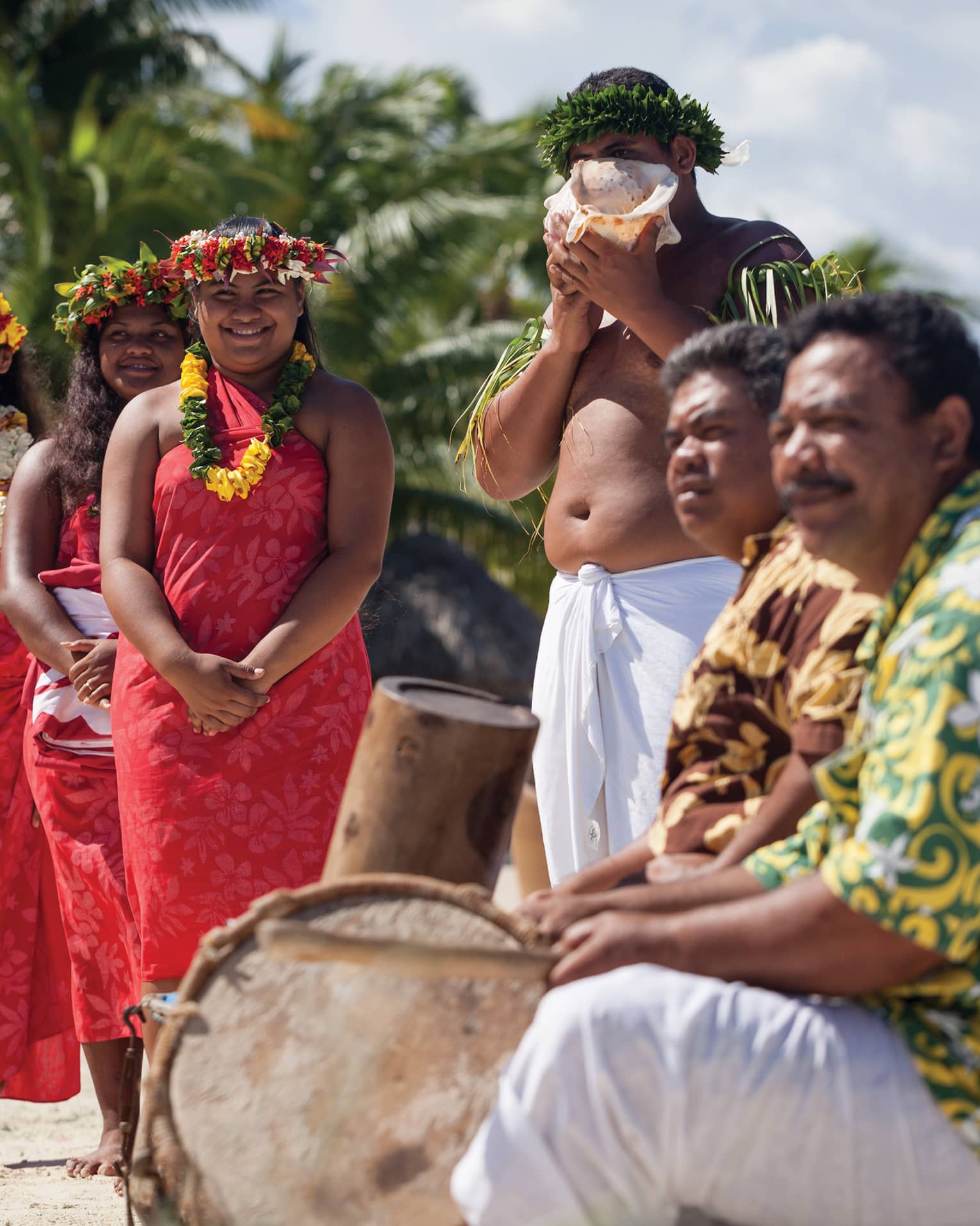 Group of people in traditional attire with leis and a conch shell, standing on a beach