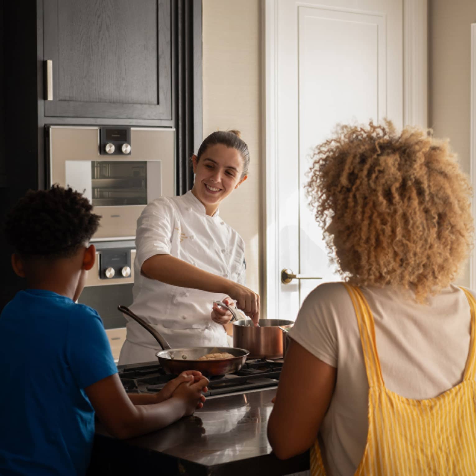 A family of four gathers around a kitchen-island cooktop watching a smiling chef prepare food in copper cookware. 