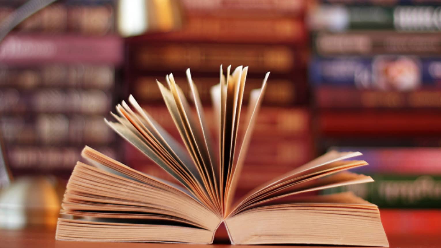 A book is opened, with pages fanned, lamp and stacks of books in background 