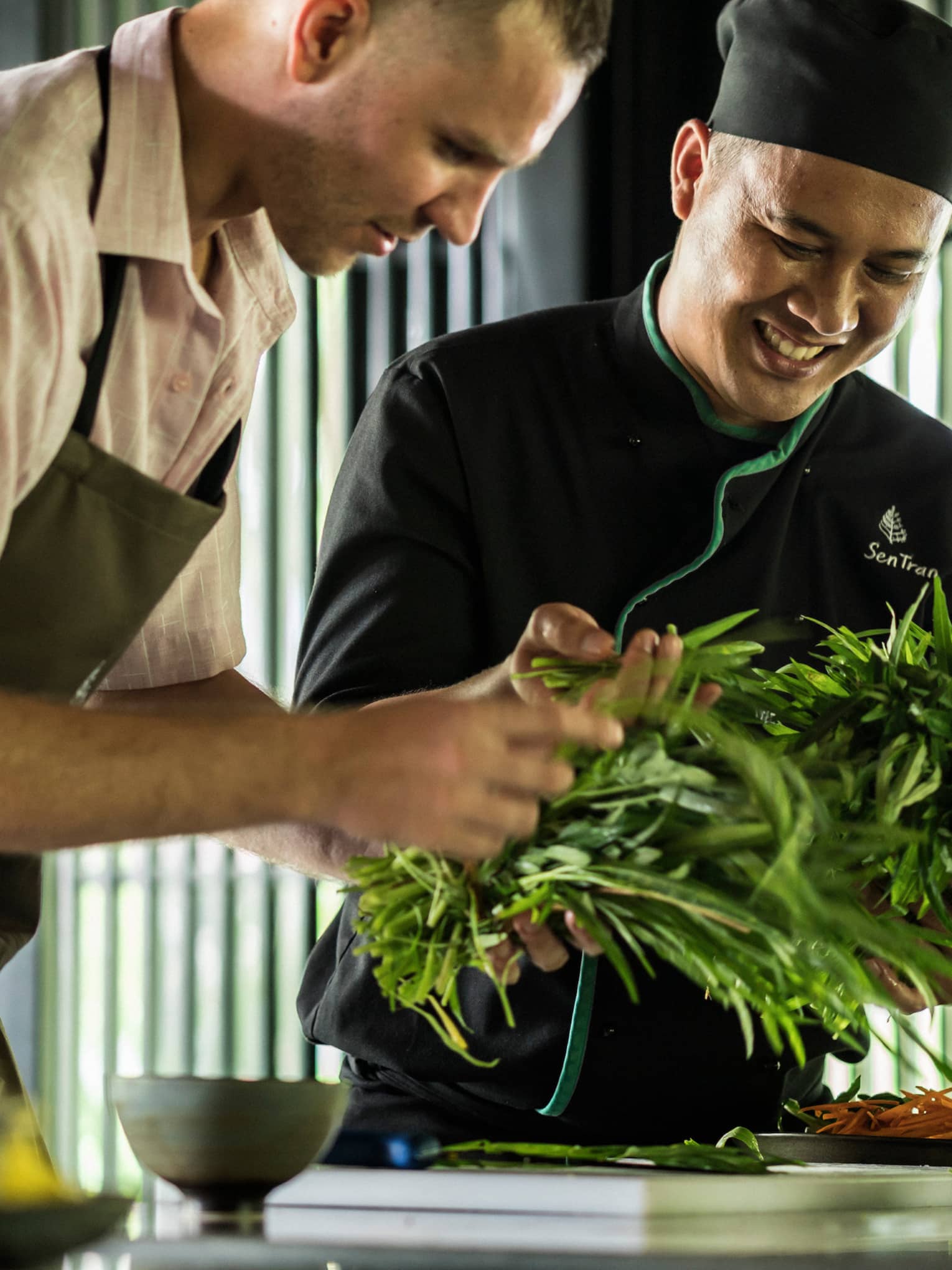 Smiling Chef Tran Van Sen holds greens, shows them to man in apron