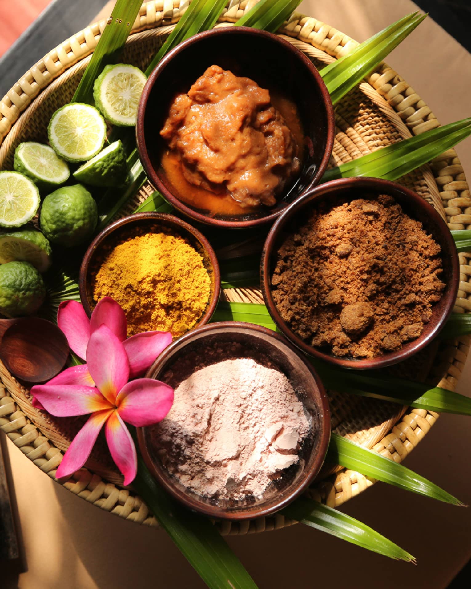 Assorted natural spa ingredients in bowls with lime, assorted spices and flower on a woven tray