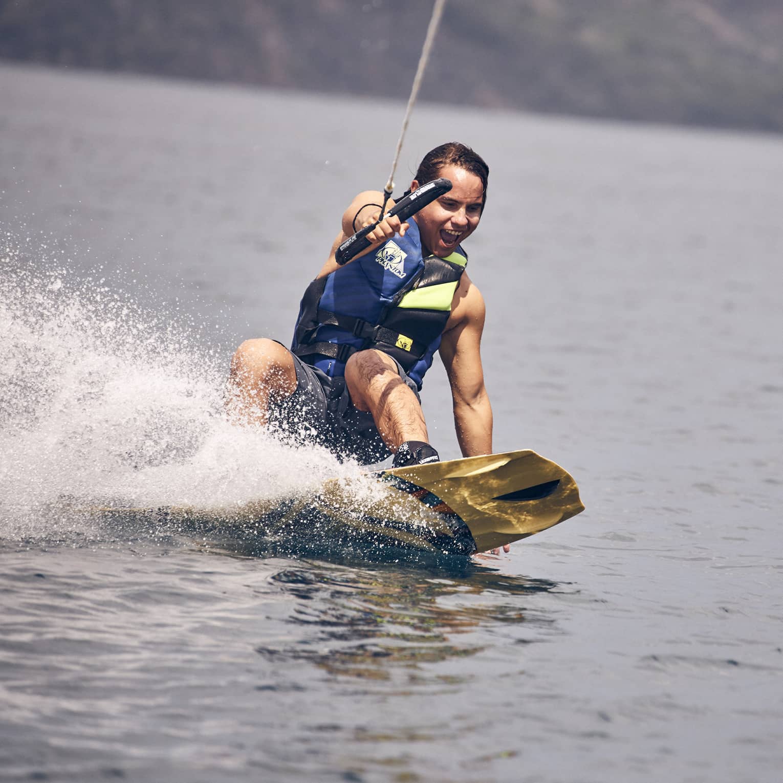 Young man wearing a blue and green life vest and blue swim trunks wakeboarding through the water
