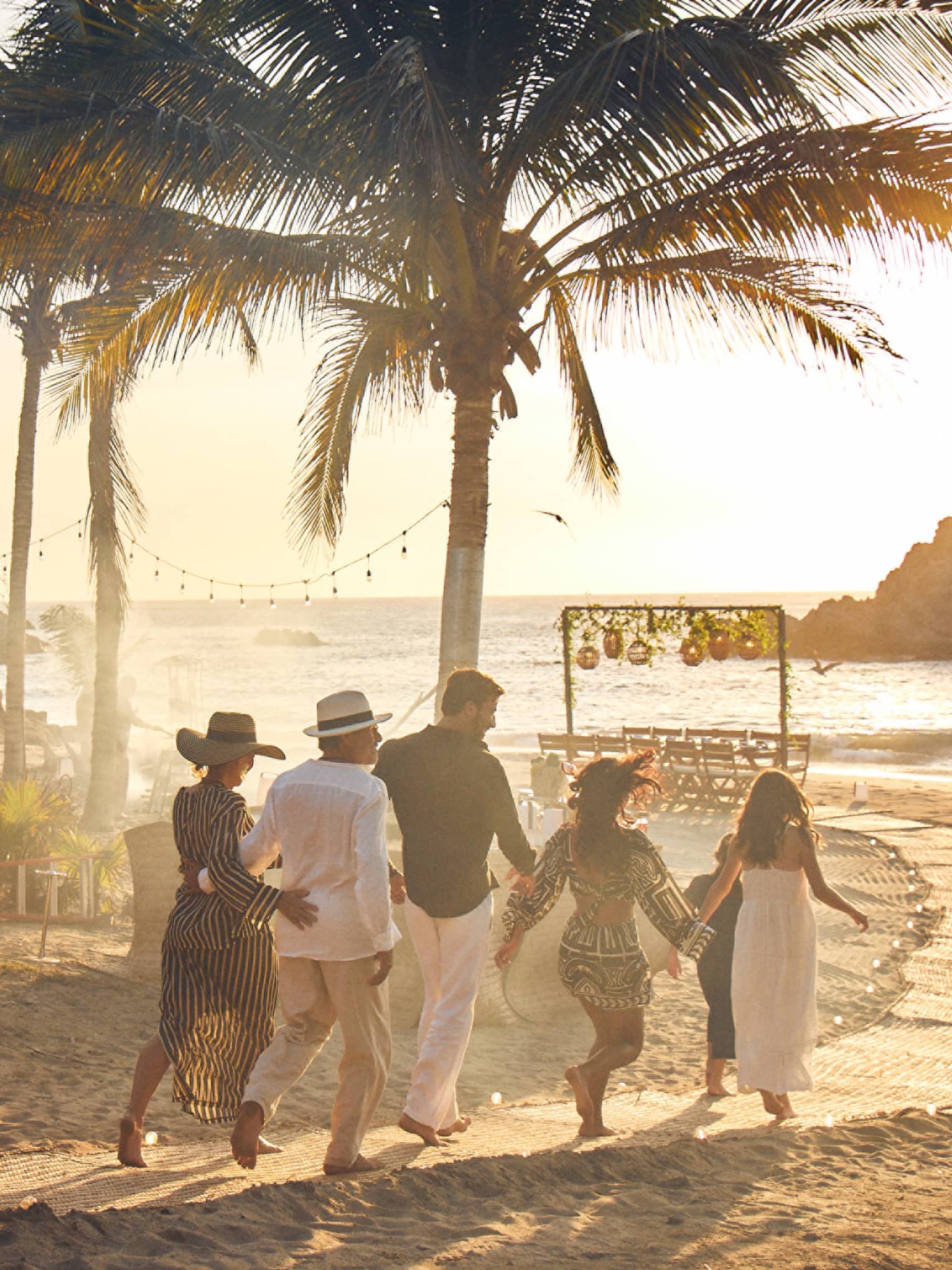 A group of adults walks down a palm-lined beach path towards a table by the ocean as the sun sets behind a rocky outcrop.