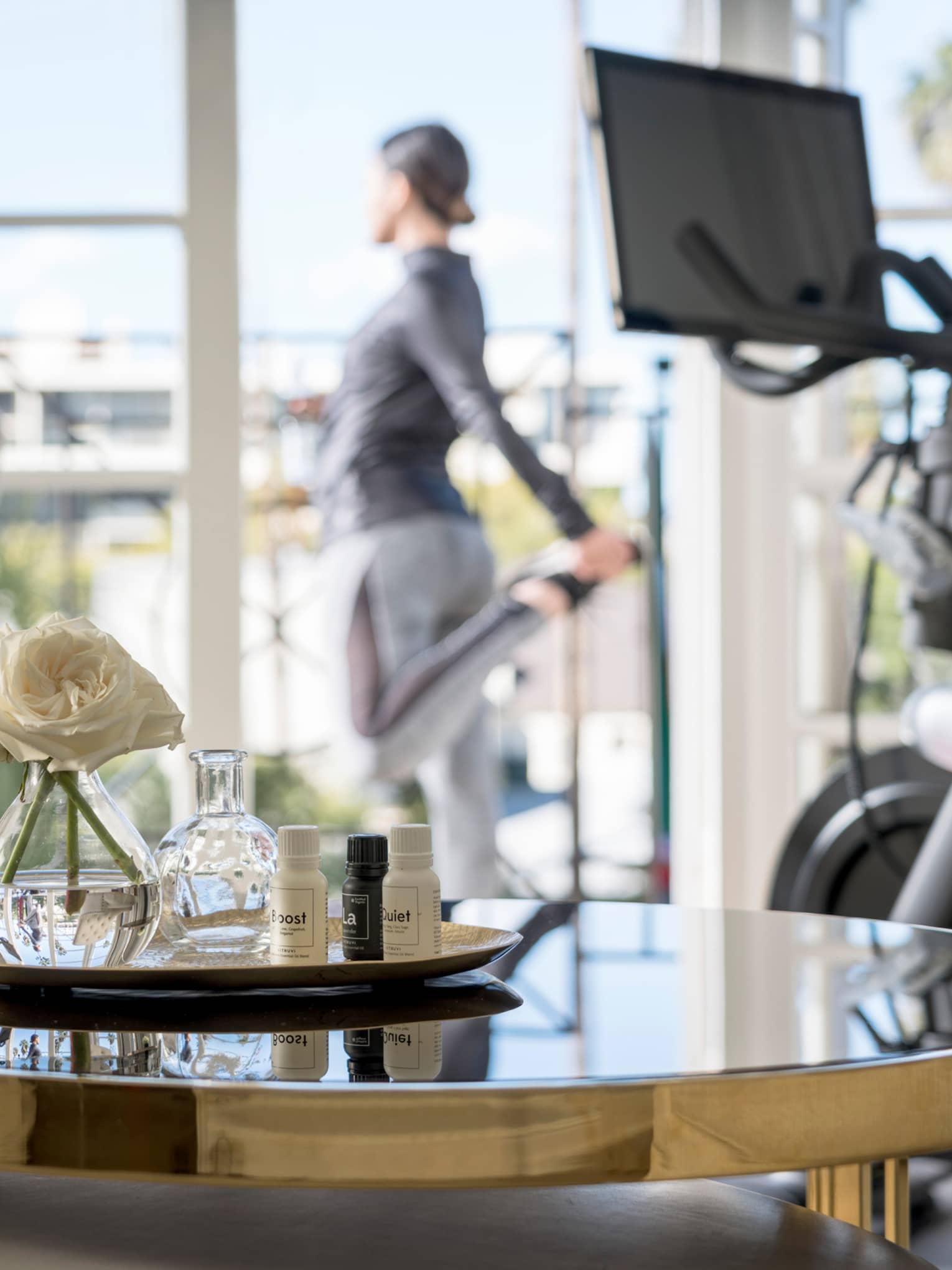 Spa tray with fresh white flowers on table by treadmill, woman stretching at balcony door