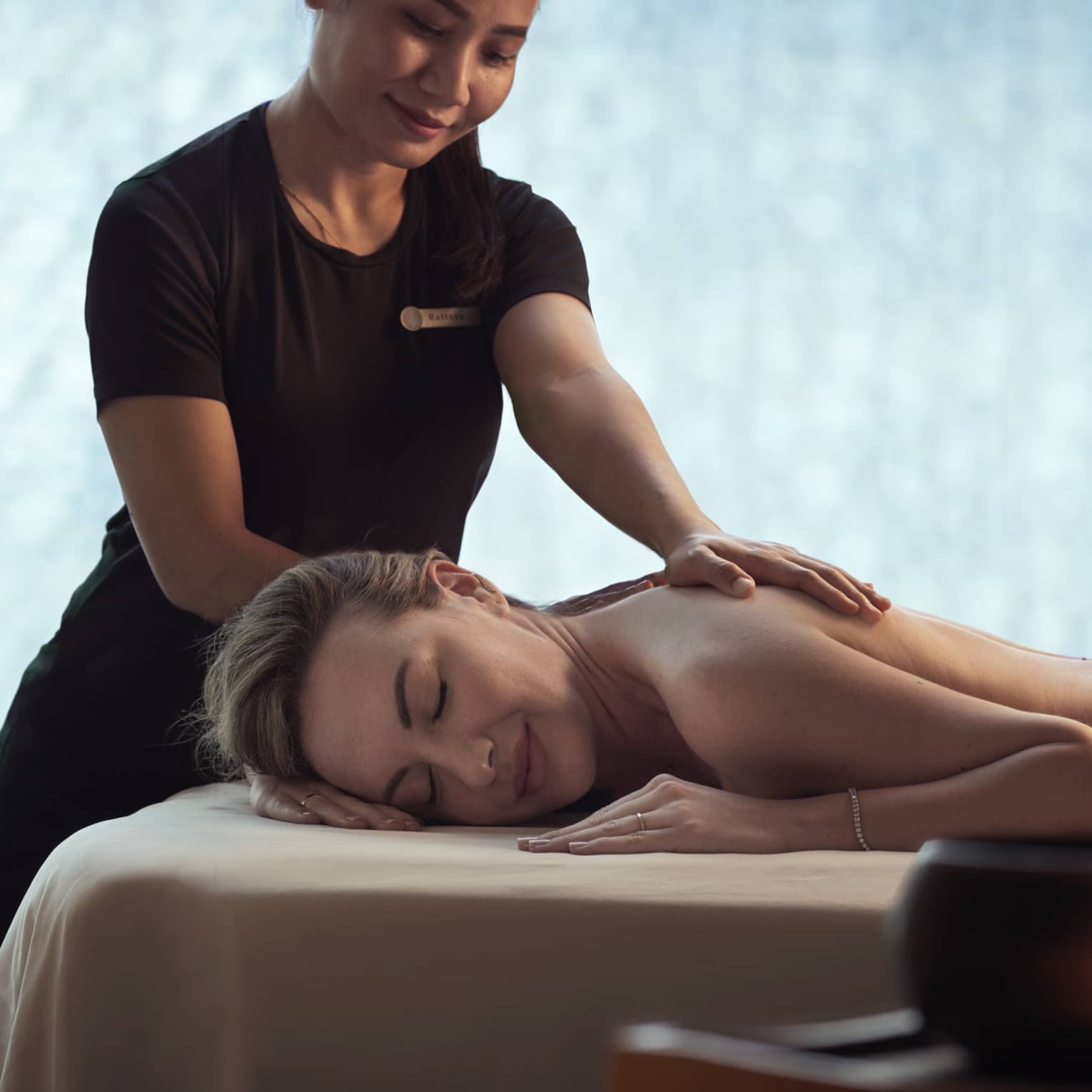 Spa staff massages woman's shoulders as she lays on bed in dark room