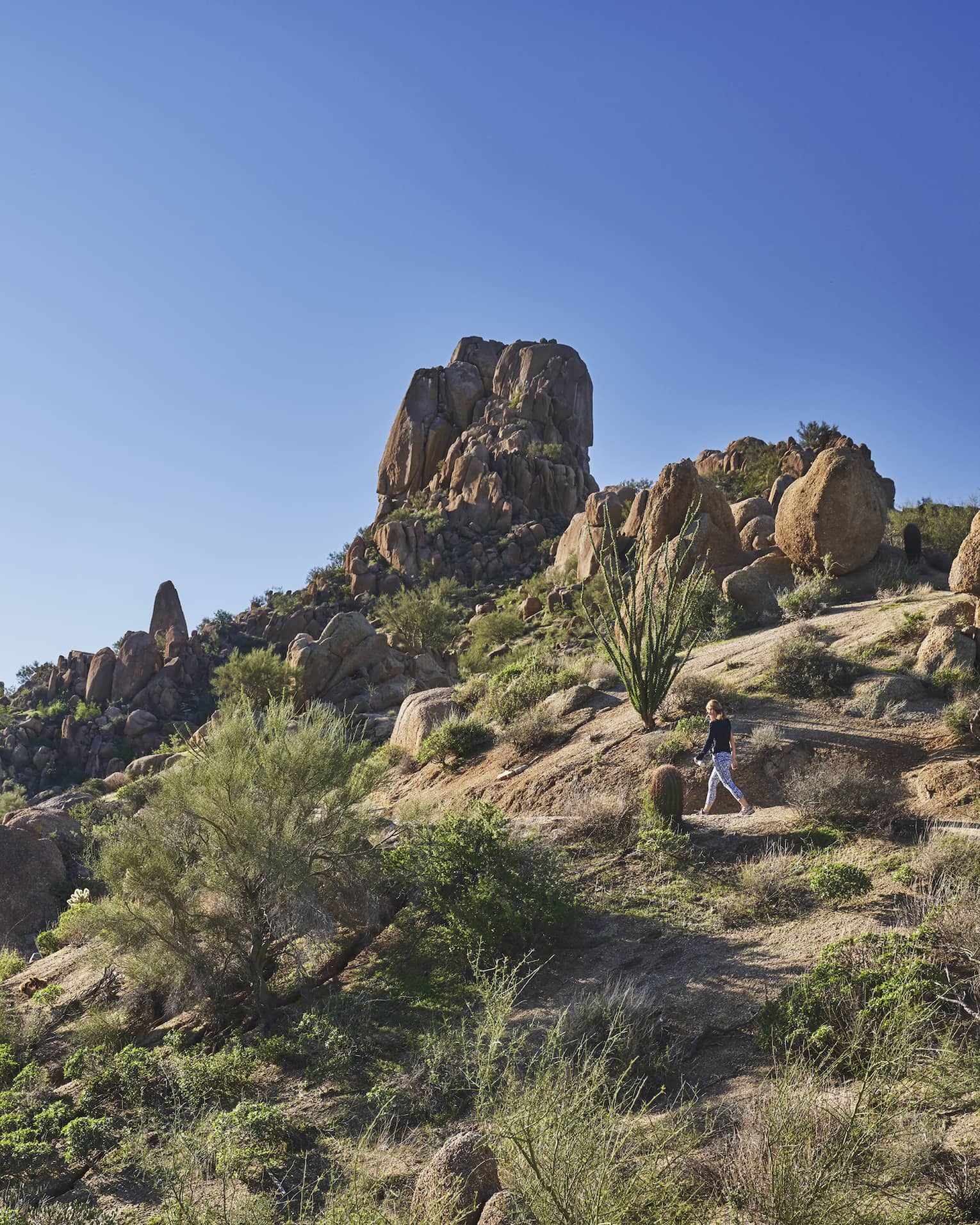 Woman walks along trail on rocky desert mountain under blue sky