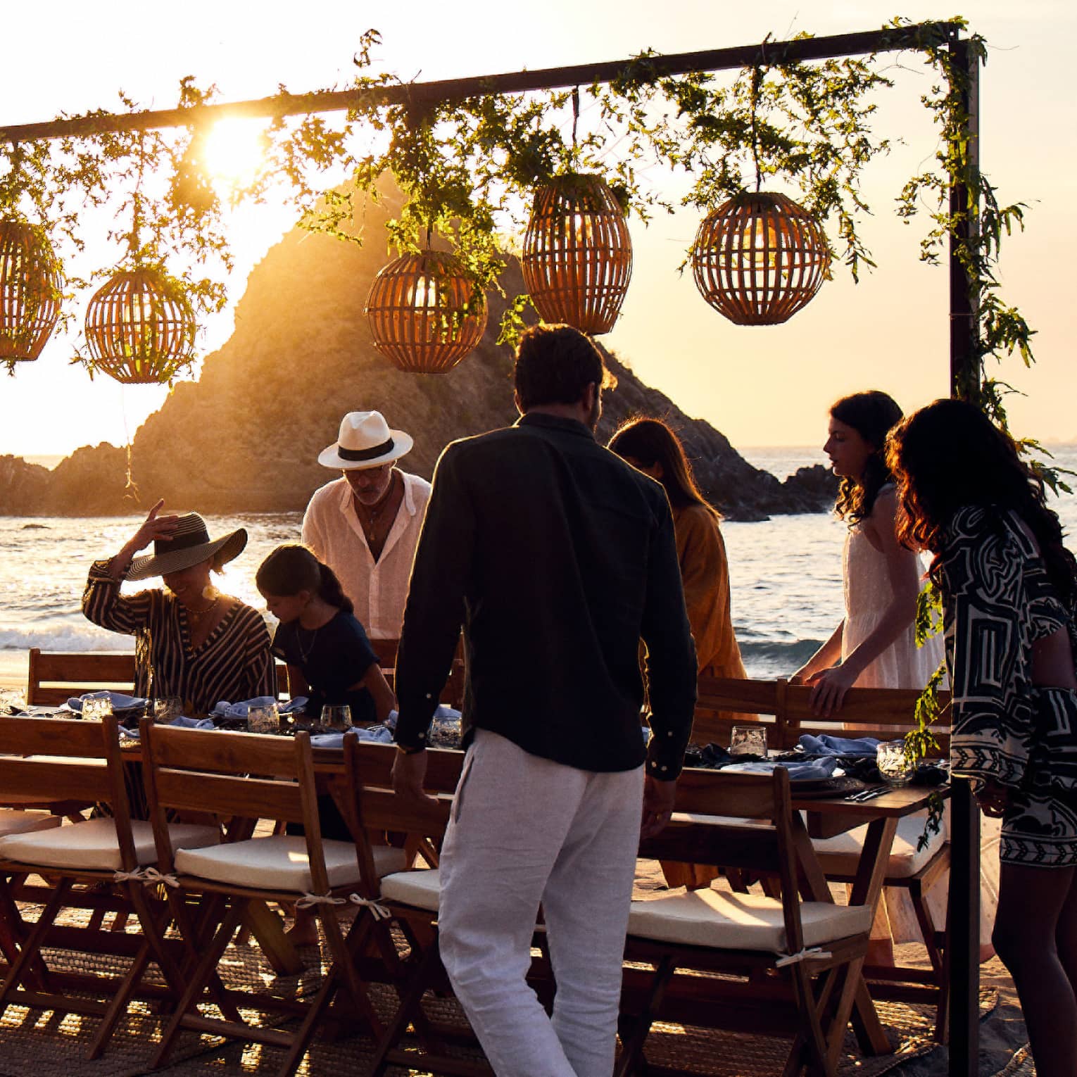 A group settles into cushioned chairs at a beachside table at dusk, lanterns and vines hanging from an attached beam above.