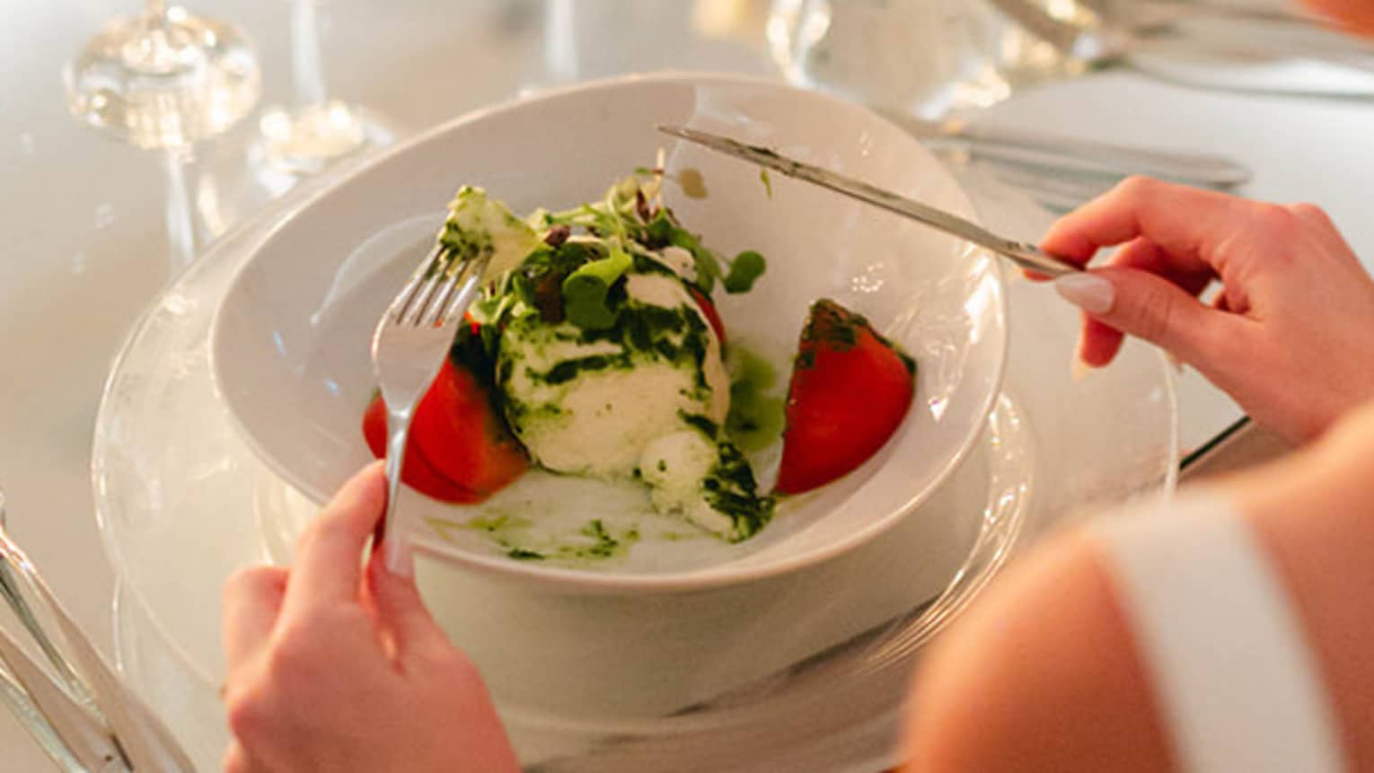 Person in bridal gown sits at round table set for dinner, fork and knife posed to dig into a meal