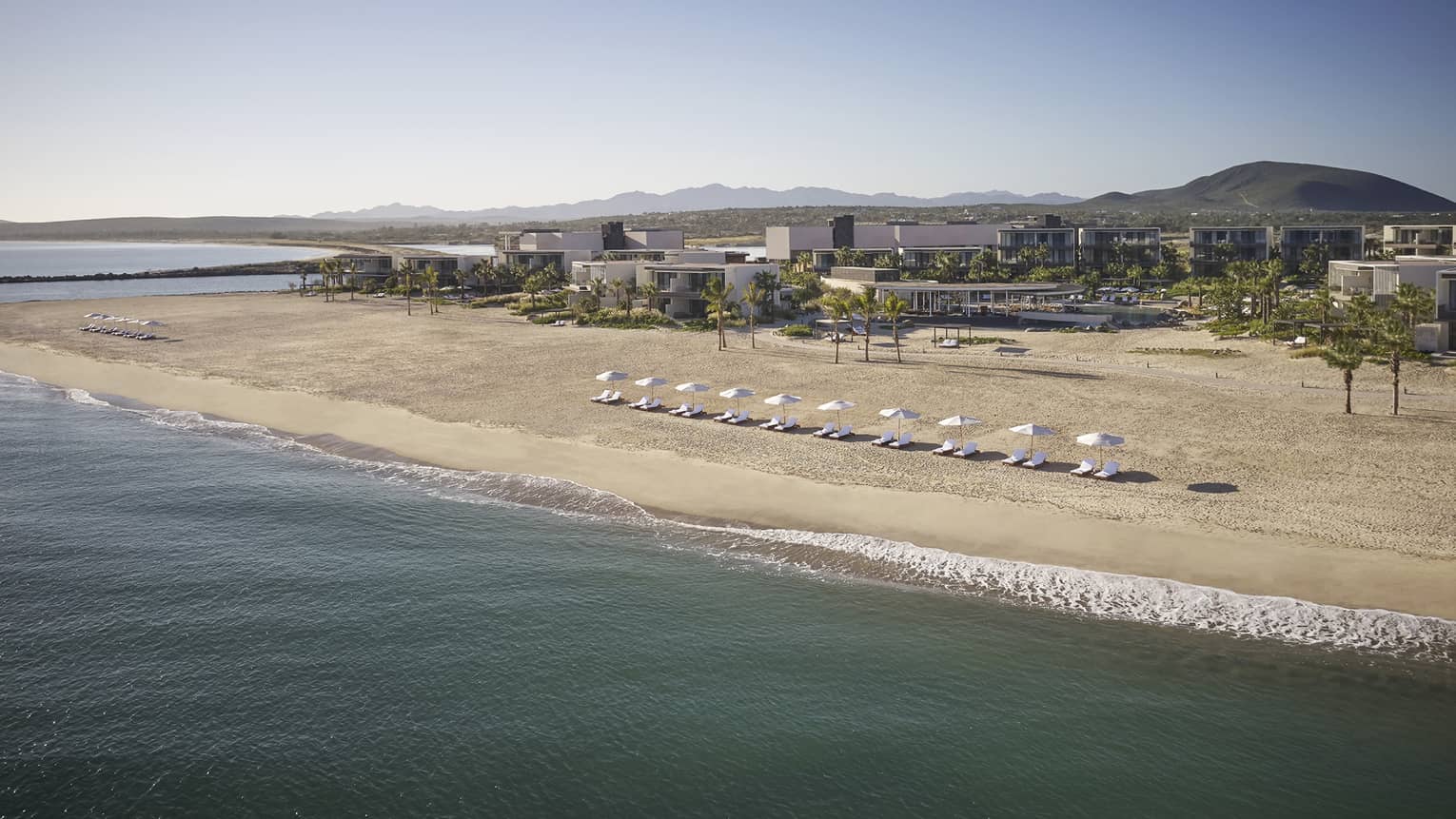 The shore of a beach with umbrellas and lounge chairs near a large building.
