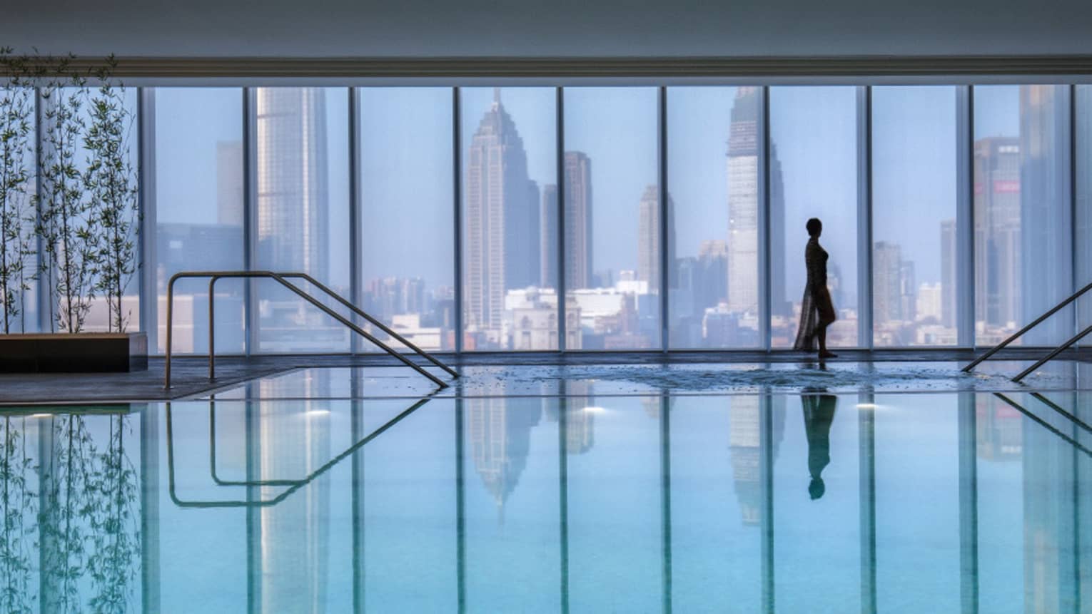 Silhouette of woman walking by blue indoor swimming pool, floor-to-ceiling window