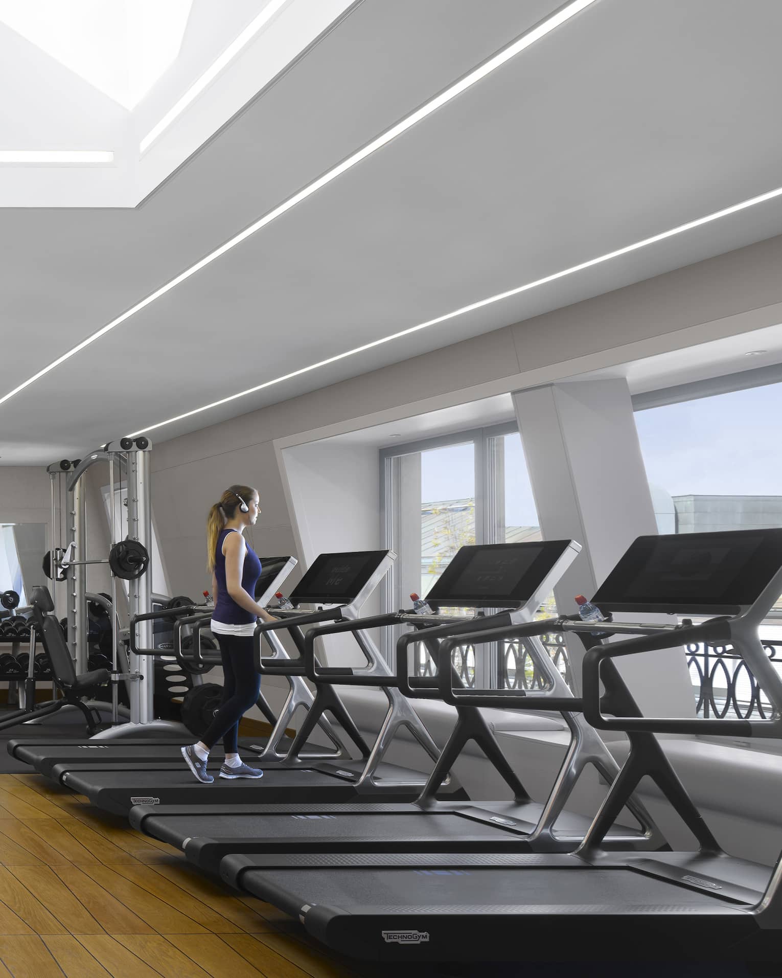 A guest walks on one of a bank of treadmills facing windows in a bright, modern gym with a striking geometric wood floor.