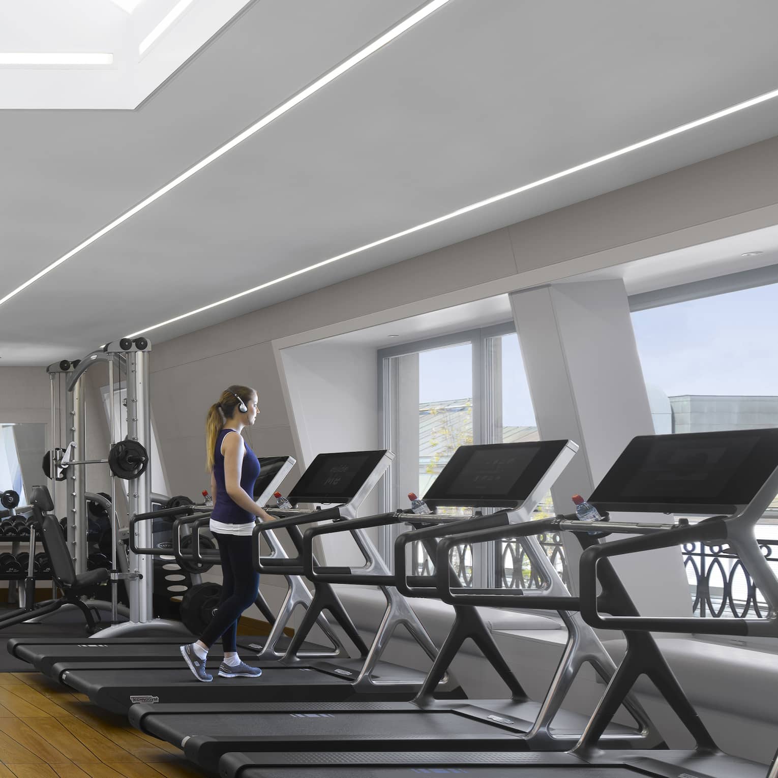 A guest walks on one of a bank of treadmills facing windows in a bright, modern gym with a striking geometric wood floor.