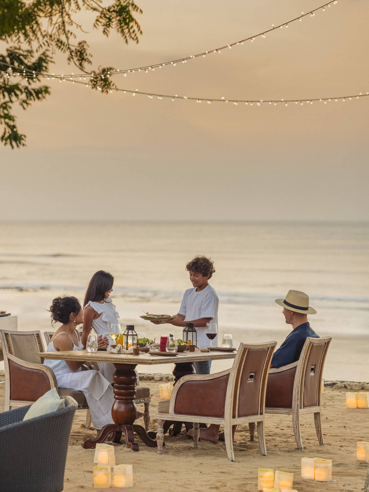 As two chefs barbecue nearby, a child brings a dish to his family’s table on a beach dotted with paper lanterns at dusk.