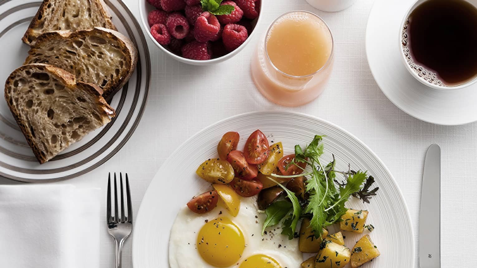 Breakfast plate with sunny-side-up eggs, cherry tomatoes, greens, and roasted potatoes, alongside toast, raspberries, grapefruit juice and coffee.