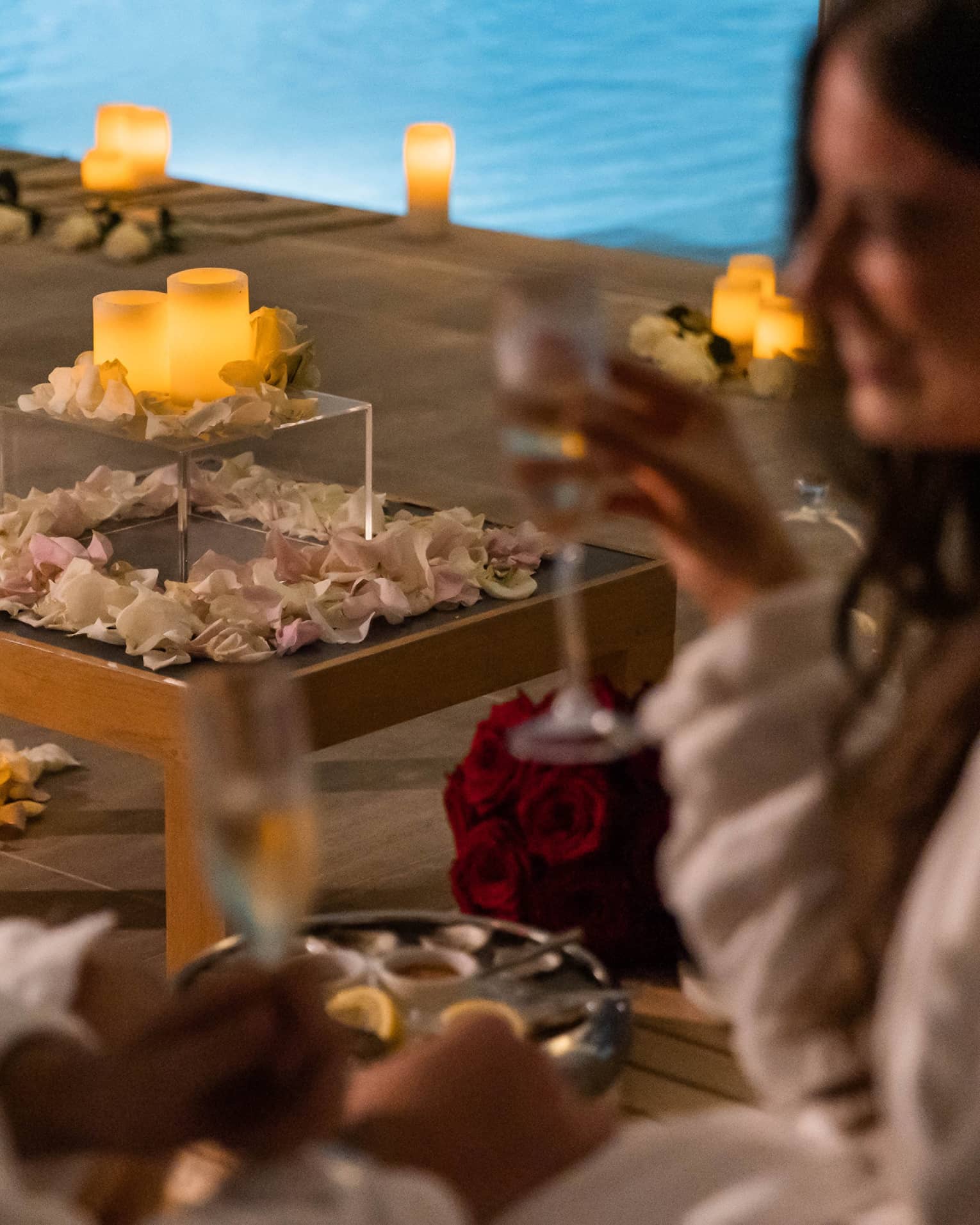A couple with wine in hand near a pool surrounded by flowers petals and candles.