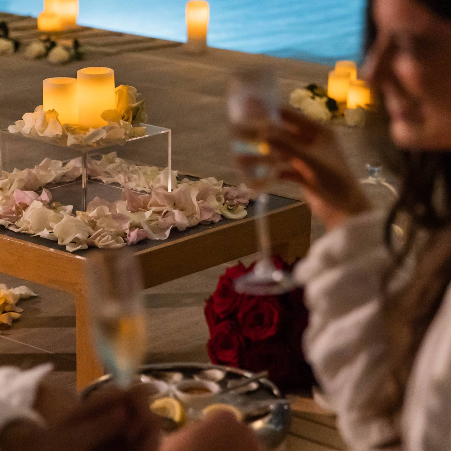 A couple with wine in hand near a pool surrounded by flowers petals and candles.