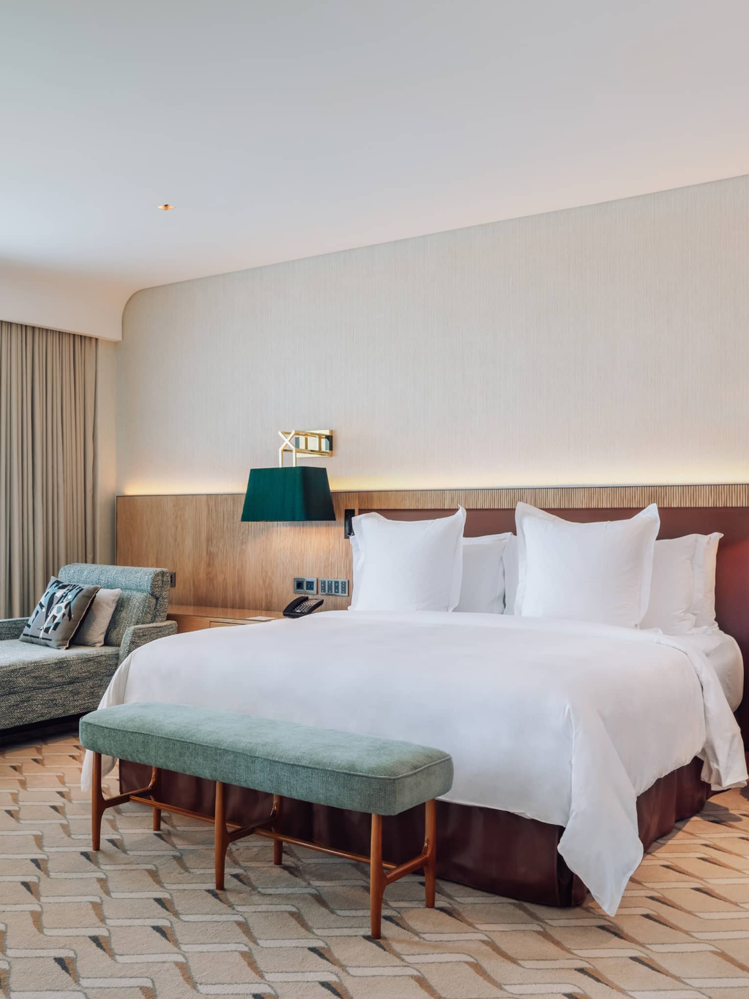 Modern hotel room with a large bed, chaise lounge, dining table, green chairs and floor-to-ceiling windows.