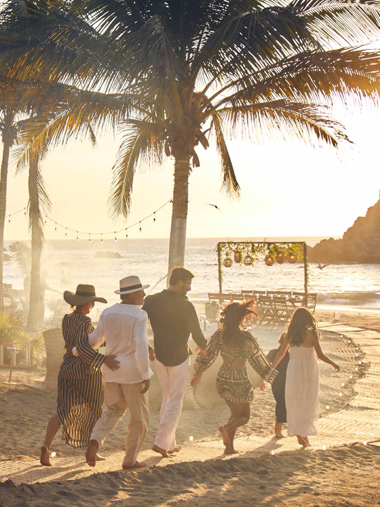 A group of adults walks down a palm-lined beach path towards a table by the ocean as the sun sets behind a rocky outcrop.