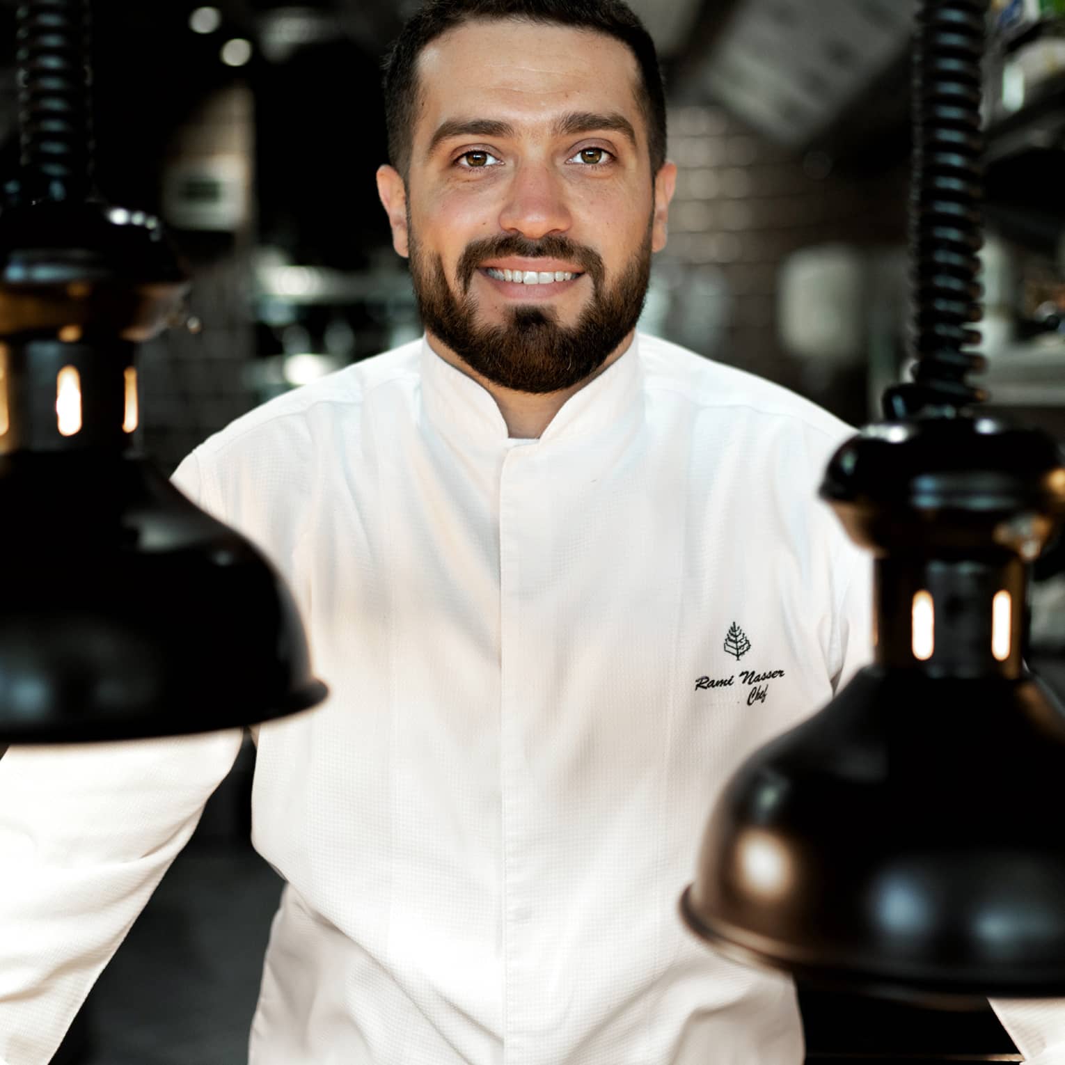 Chef Rami Nasser in his white chef's jacket smiles at the camera, flanked by black vintage pendant lamps hanging from coils.