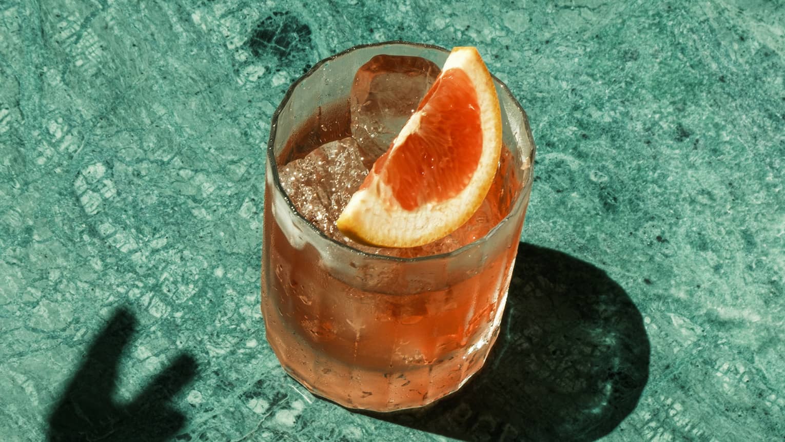 A light orange cocktail garnished with an orange segment sits on a marbled blue-green surface. There is a shadow cast next to the glass by a hand reaching toward it.