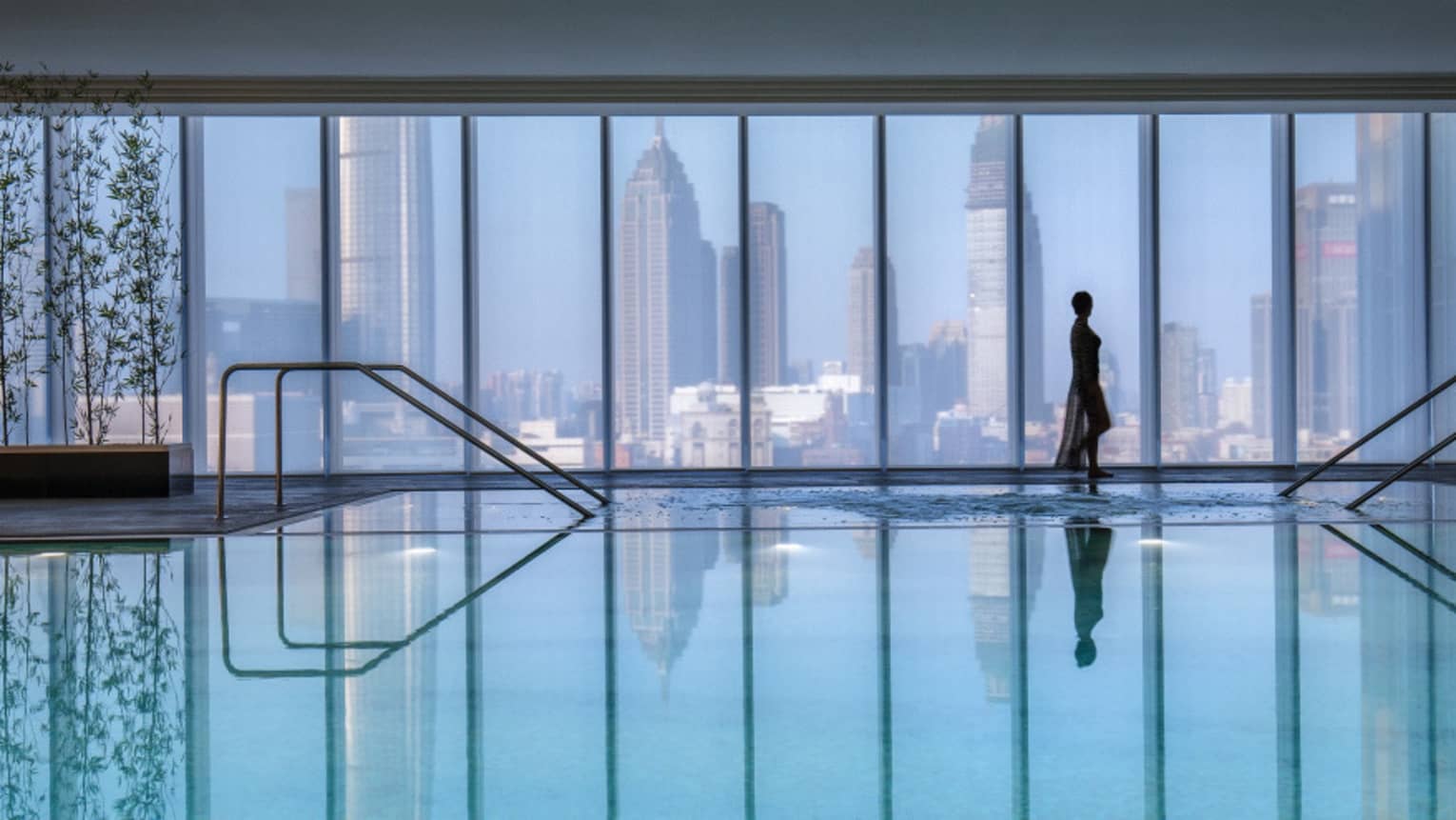 Silhouette of woman walking by blue indoor swimming pool, floor-to-ceiling window