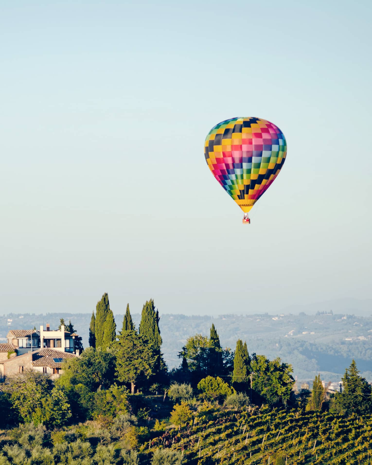 Colourful hot air balloon floating above a landscape with trees and houses, set against a clear blue sky.		
