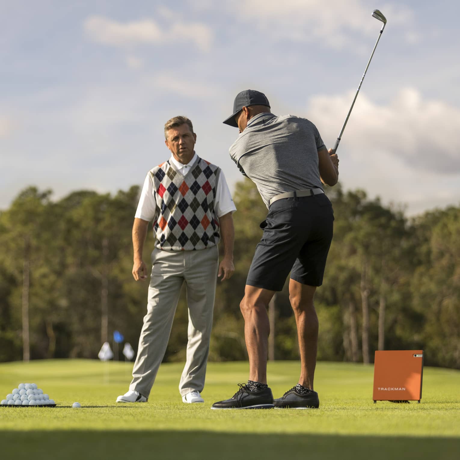 Against clear sky, one golfer looks on as another prepares to swing, a tray of balls and a stand of clubs nearby.