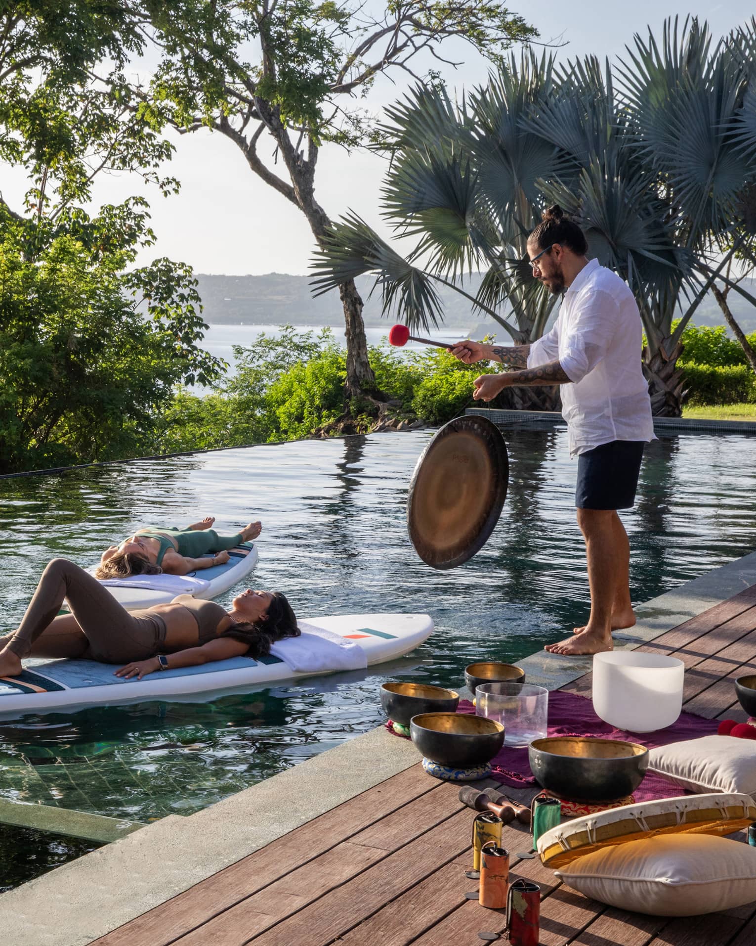 Three people float on stand-up paddleboards in a private pool while someone plays a gong on the deck