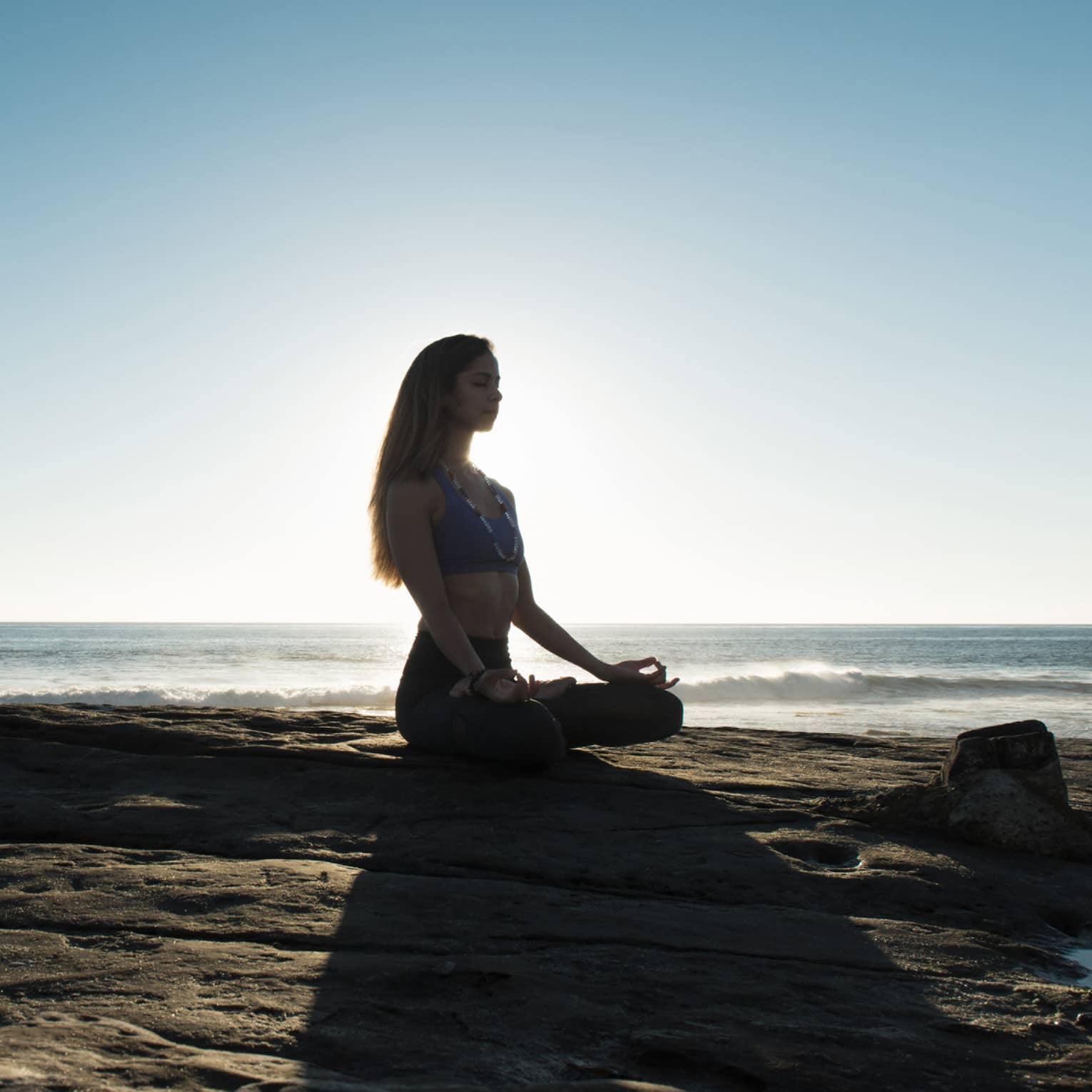 Silhouette of woman sitting cross-legged, meditating on rock by ocean
