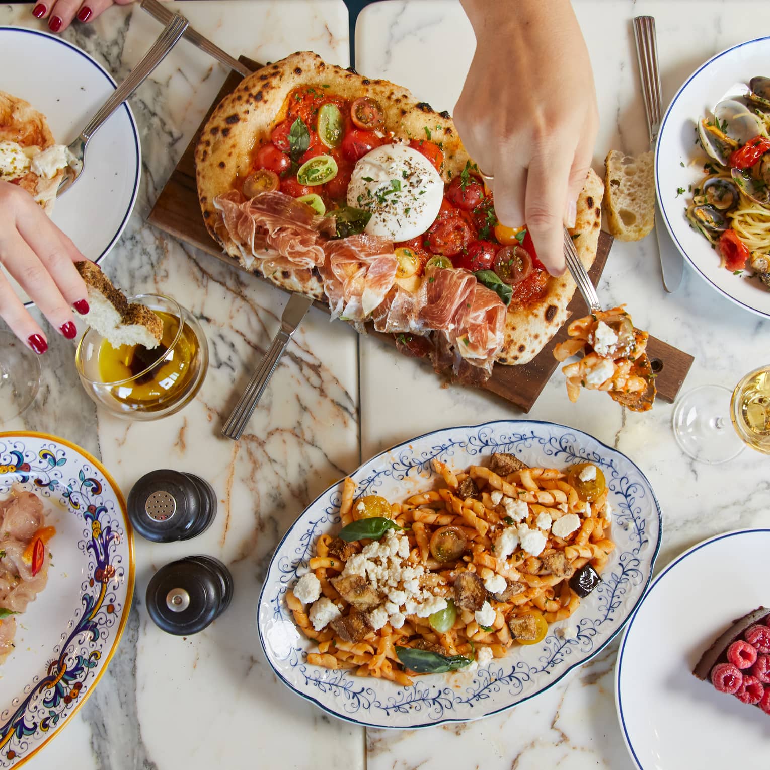 A marble table features white wine and an array of colourful Mediterranean delicacies, hands reaching in to sample the food.