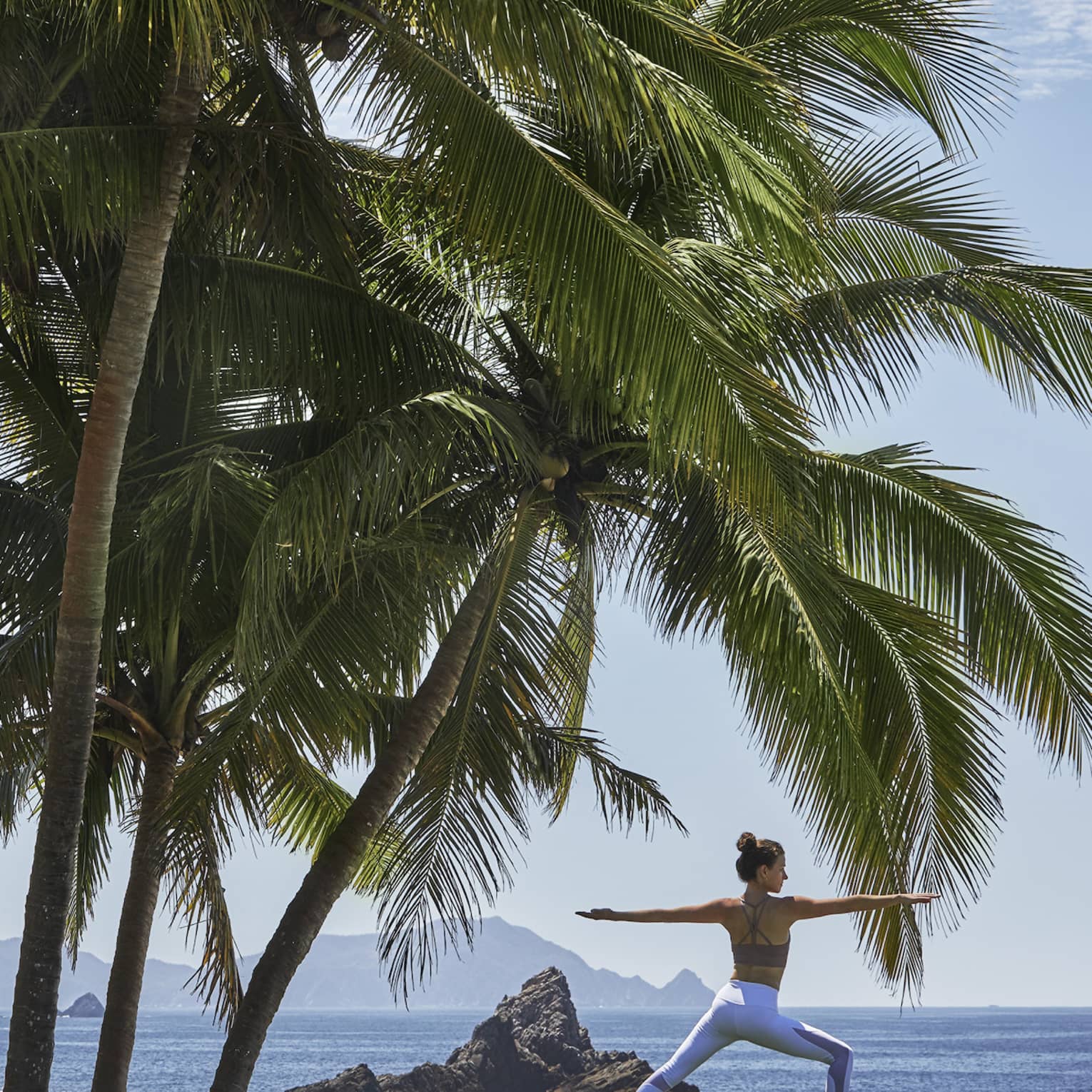 A guest in warrior two yoga pose, dwarfed by a nearby group of immense palm trees, along a sea shore dotted by rock spires. 
