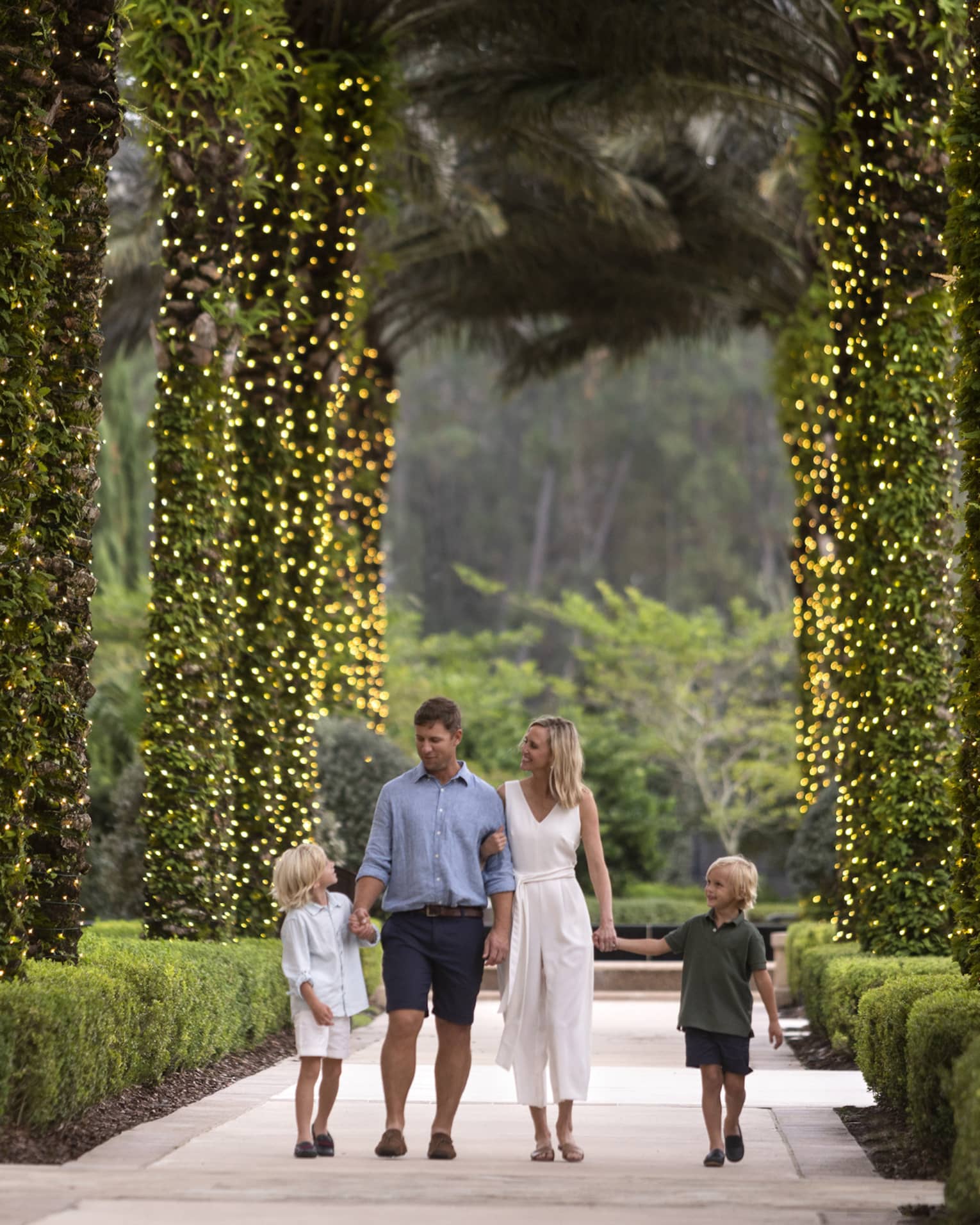 A mother, father and two sons walking along a path of palm trees wrapped in Christmas lights.