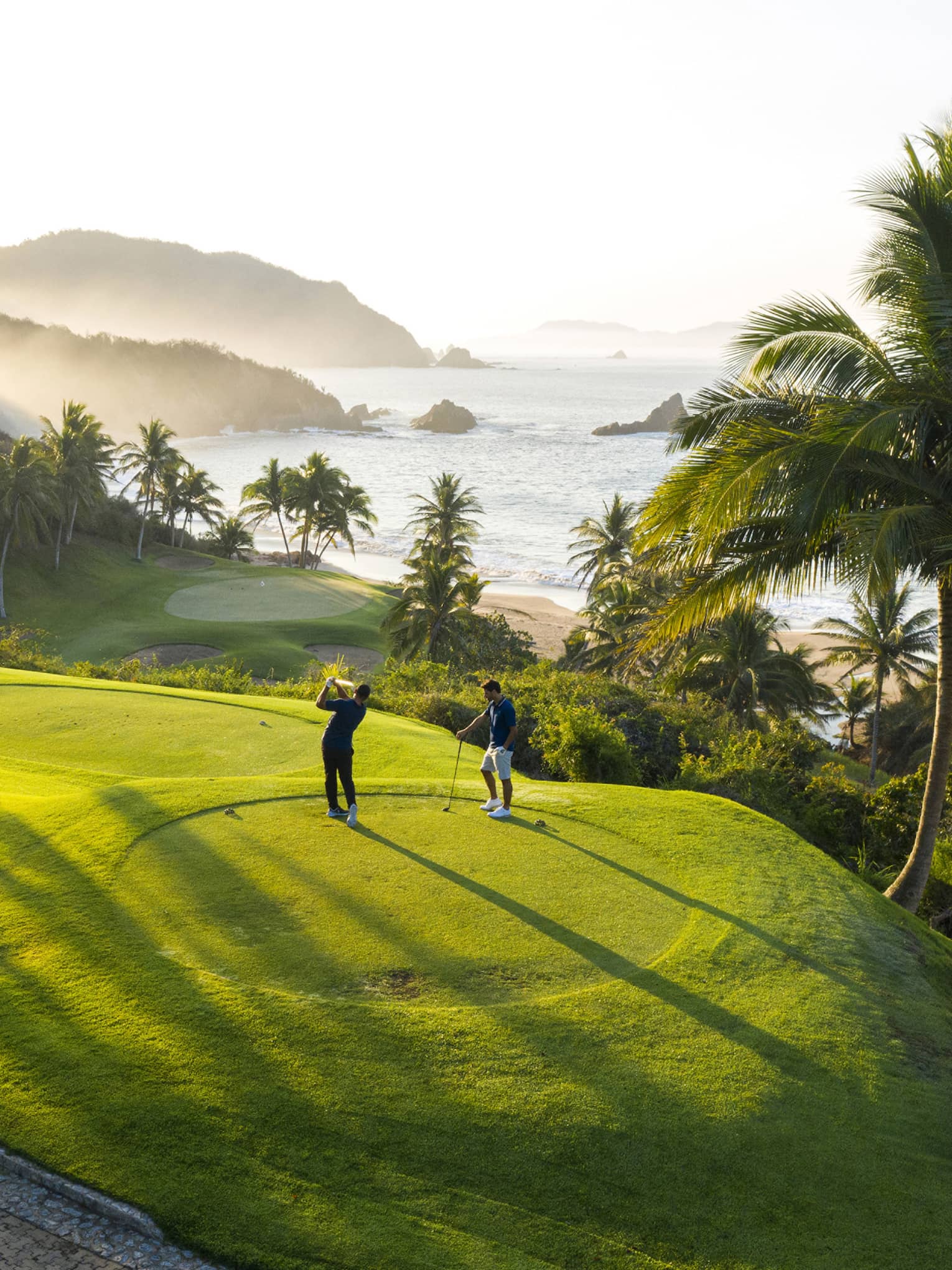 Rear view of two golfers on an elevated tee, one swinging toward the green down by the beach, misty hills and ocean beyond.