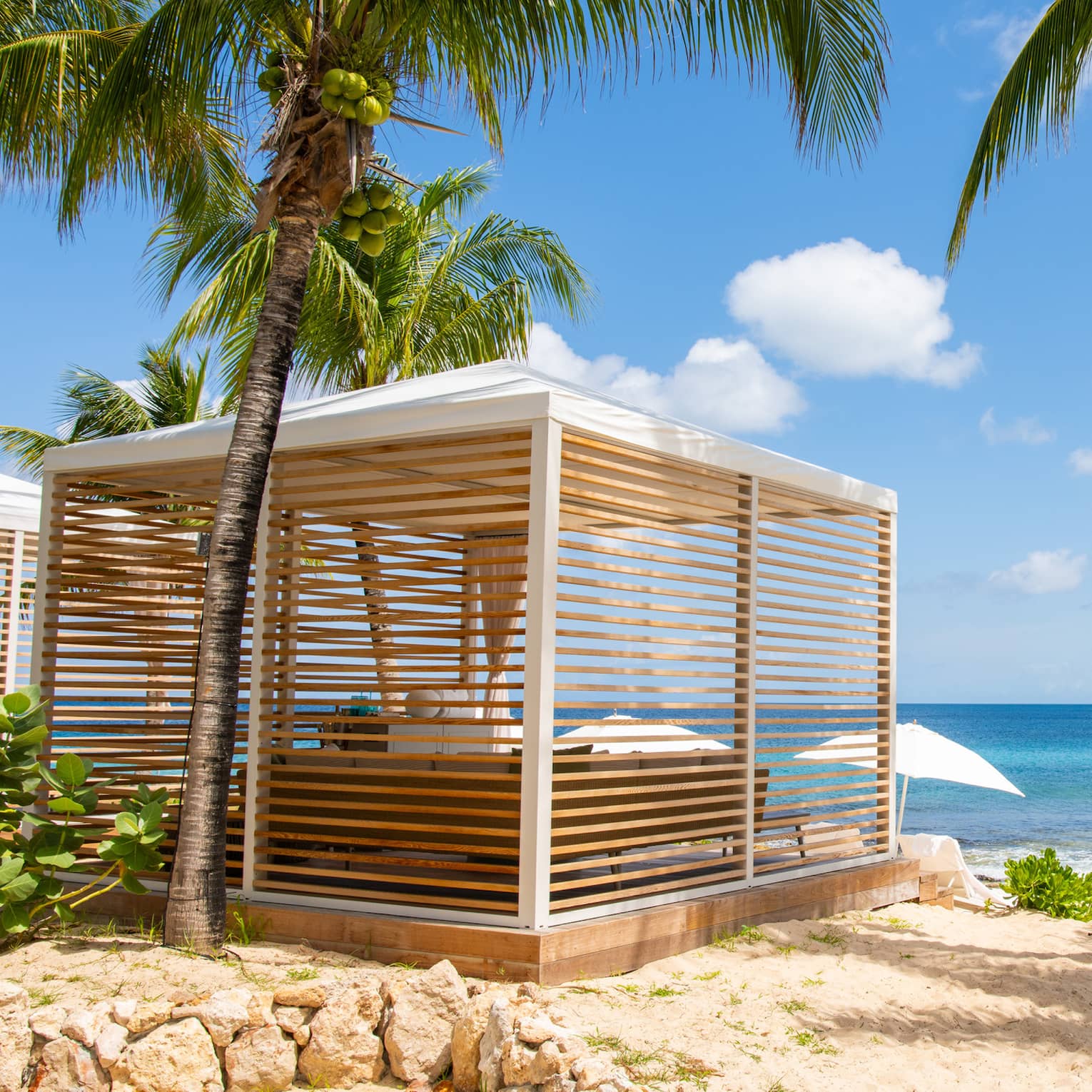 Luxury resort cabana with wooden slats surrounded by palm trees, located on a sandy beach and overlooking the ocean 