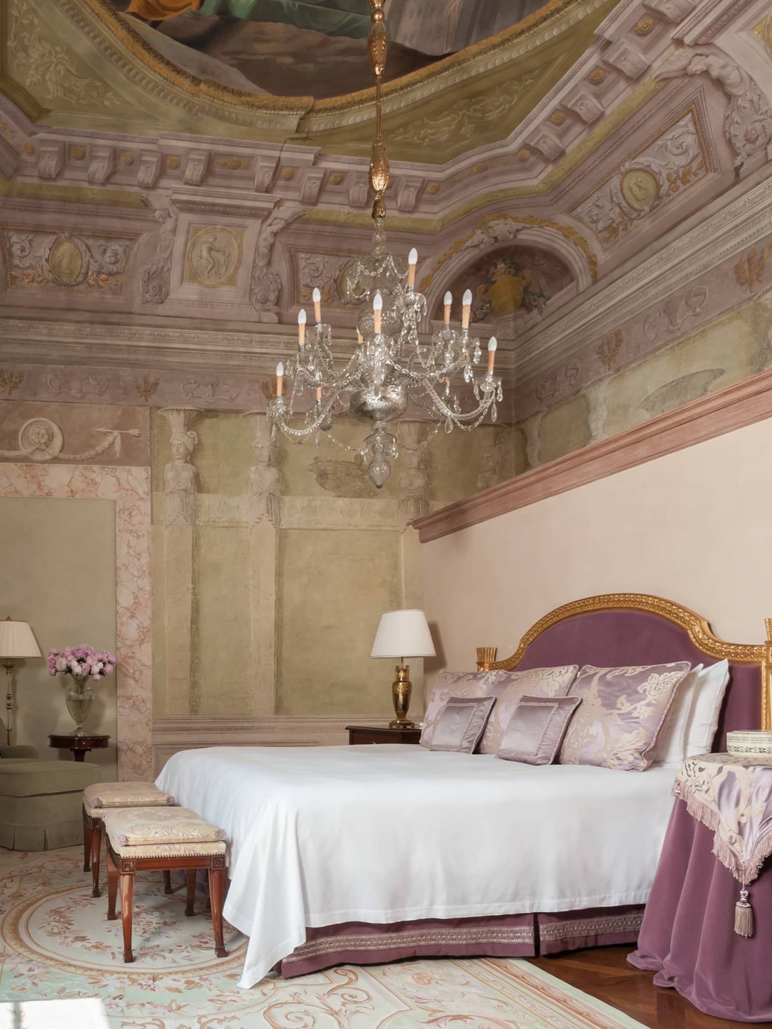 Frescoed Executive Suite bed with purple velvet headboard, nightstand with cloth under chandelier, vaulted ceiling