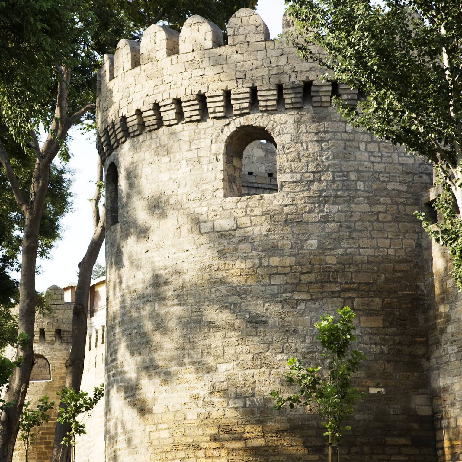 A cylindrical structure marked by loophole openings and grey stone exterior: part of a crenelated tower surrounded by trees. 