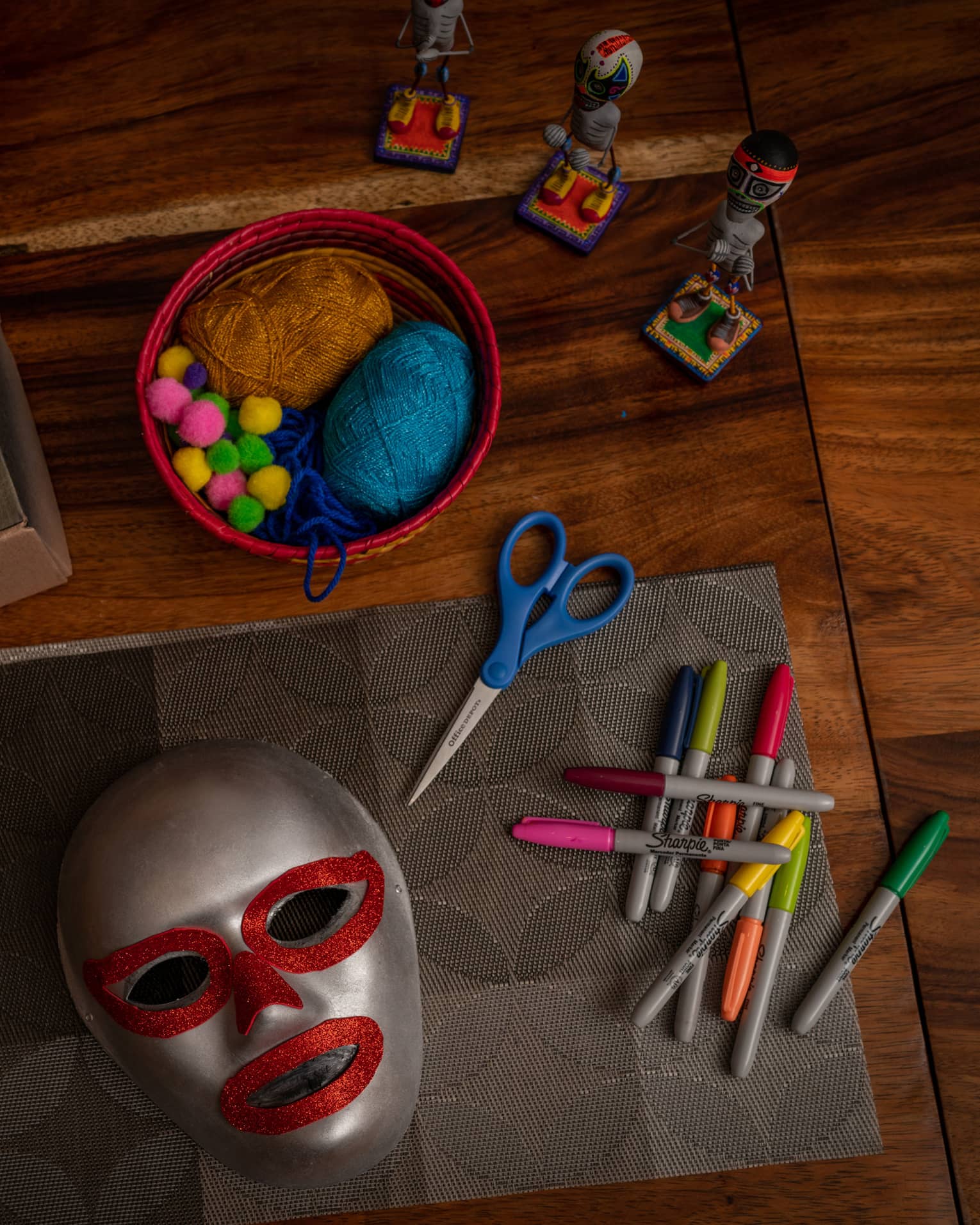 Atop a wooden surface, small figurines, masks and mask-making supplies: scissors, colourful markers, pompoms and yarn.