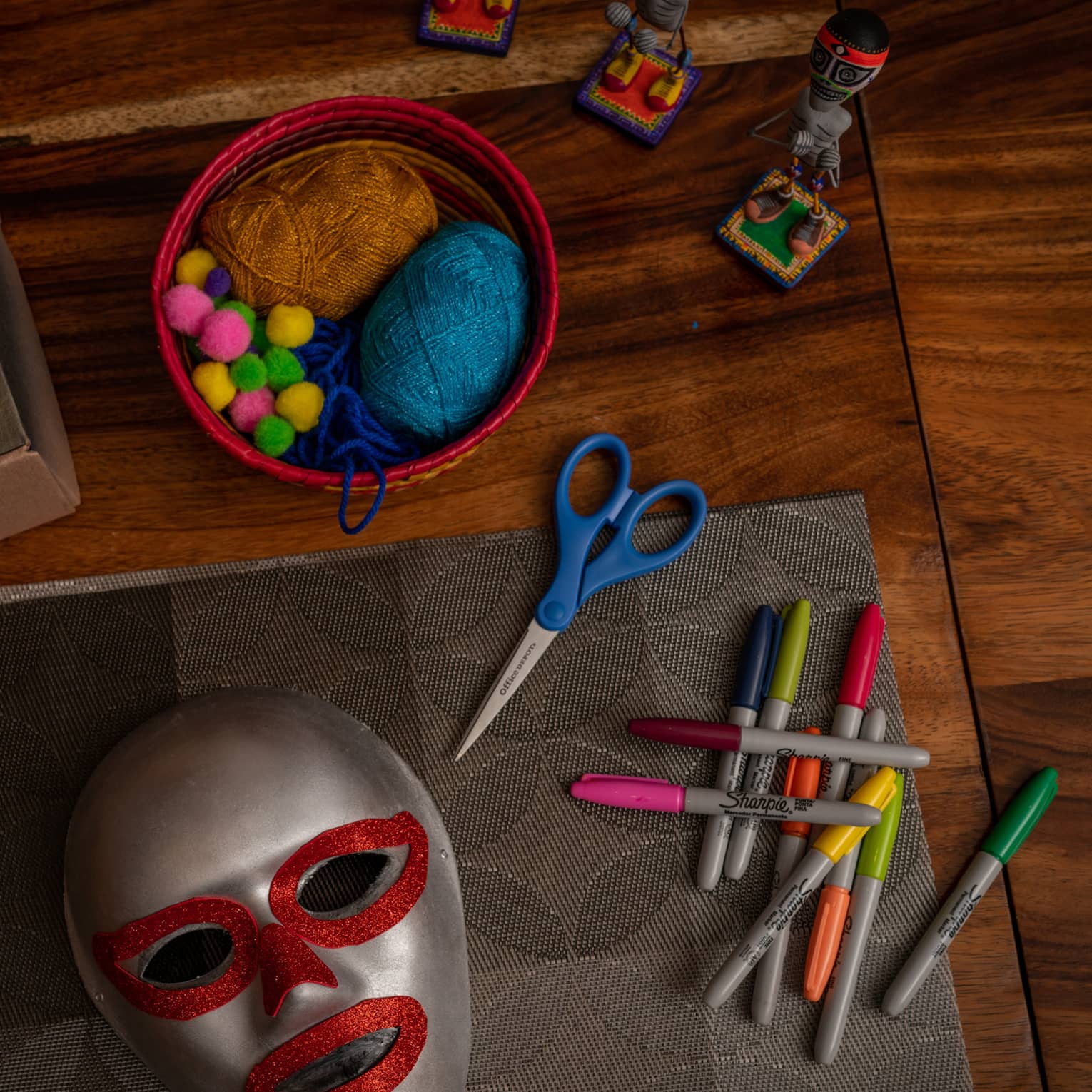 Atop a wooden surface, small figurines, masks and mask-making supplies: scissors, colourful markers, pompoms and yarn.