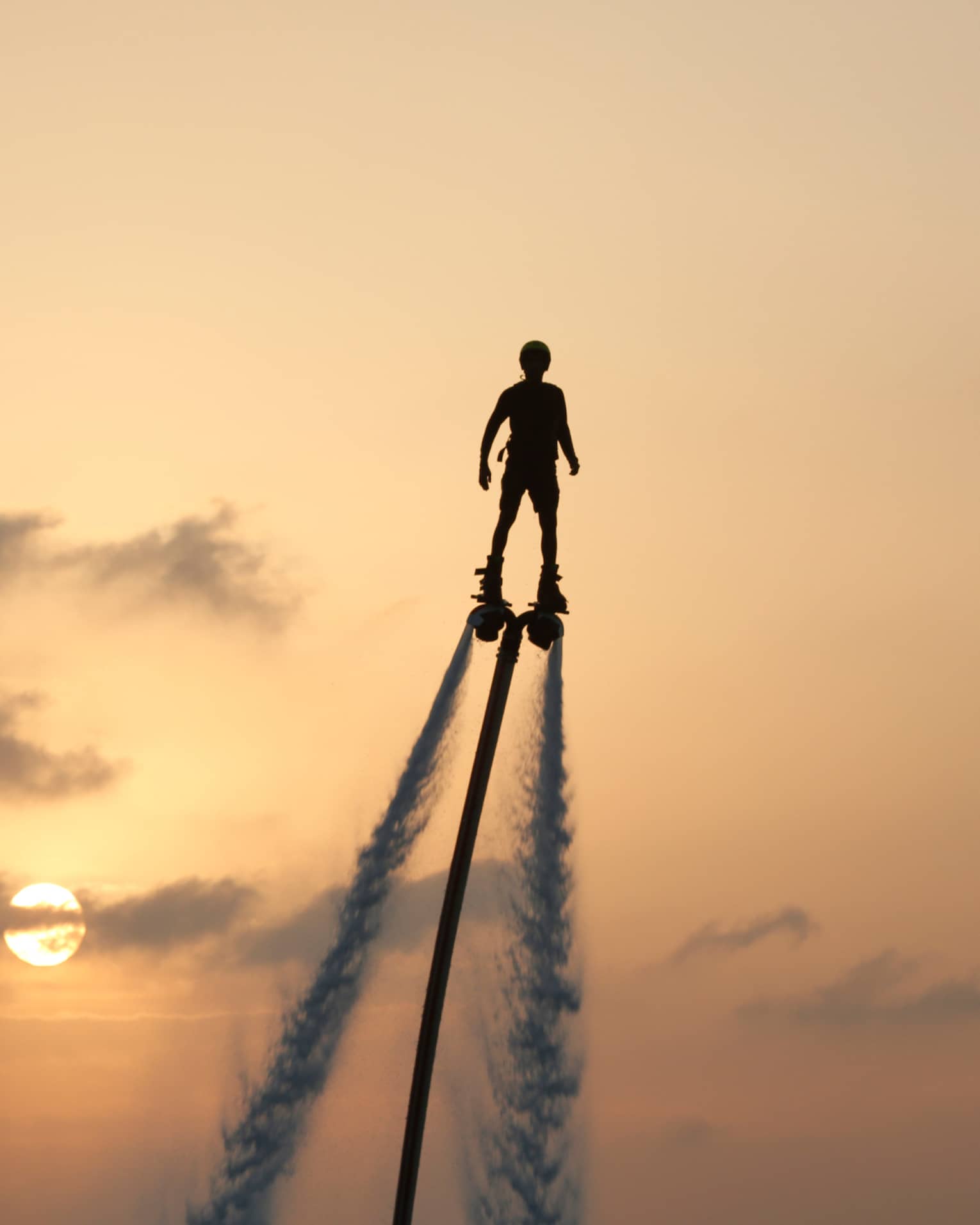 Silhouette of person soaring high above ocean on jet pack, water trails below, against sunset
