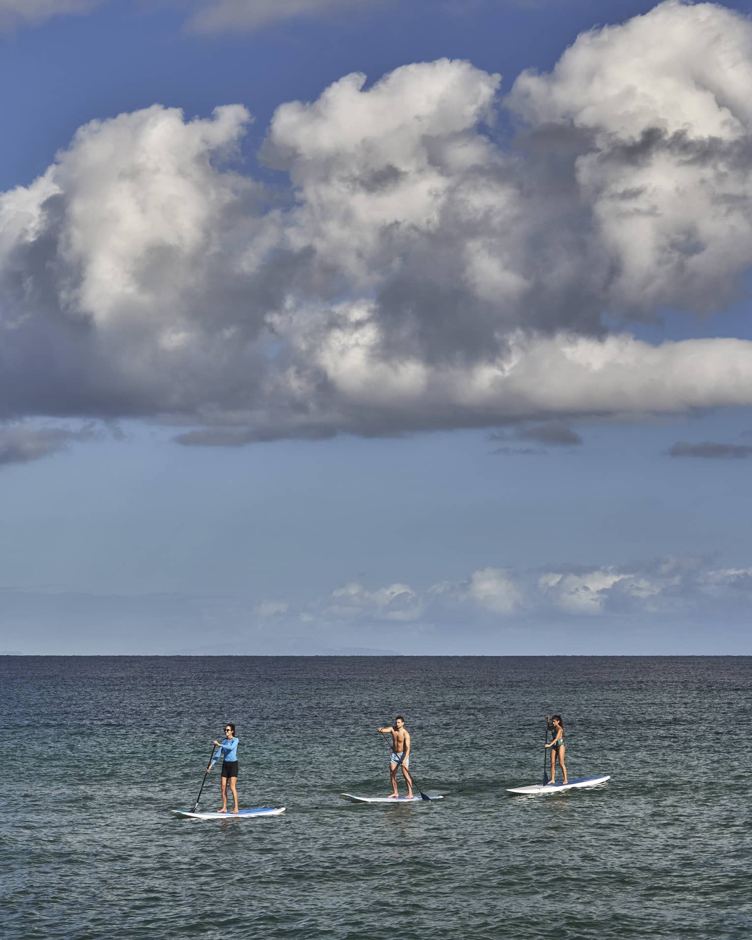 Three people on stand-up paddleboards glide on open, calm water under an expanse of blue sky and a large dramatic cloud.