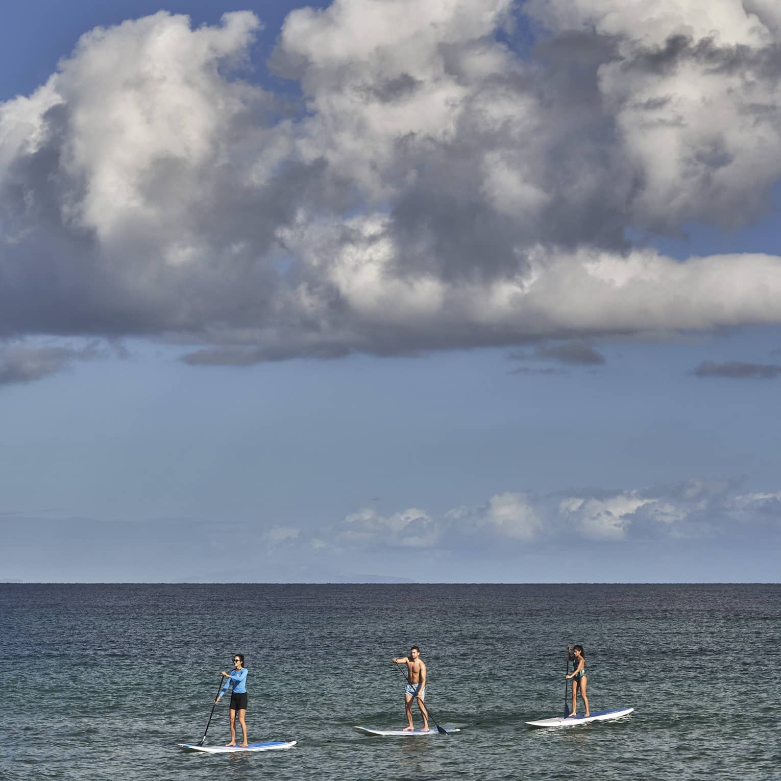 Three people on stand-up paddleboards glide on open, calm water under an expanse of blue sky and a large dramatic cloud.