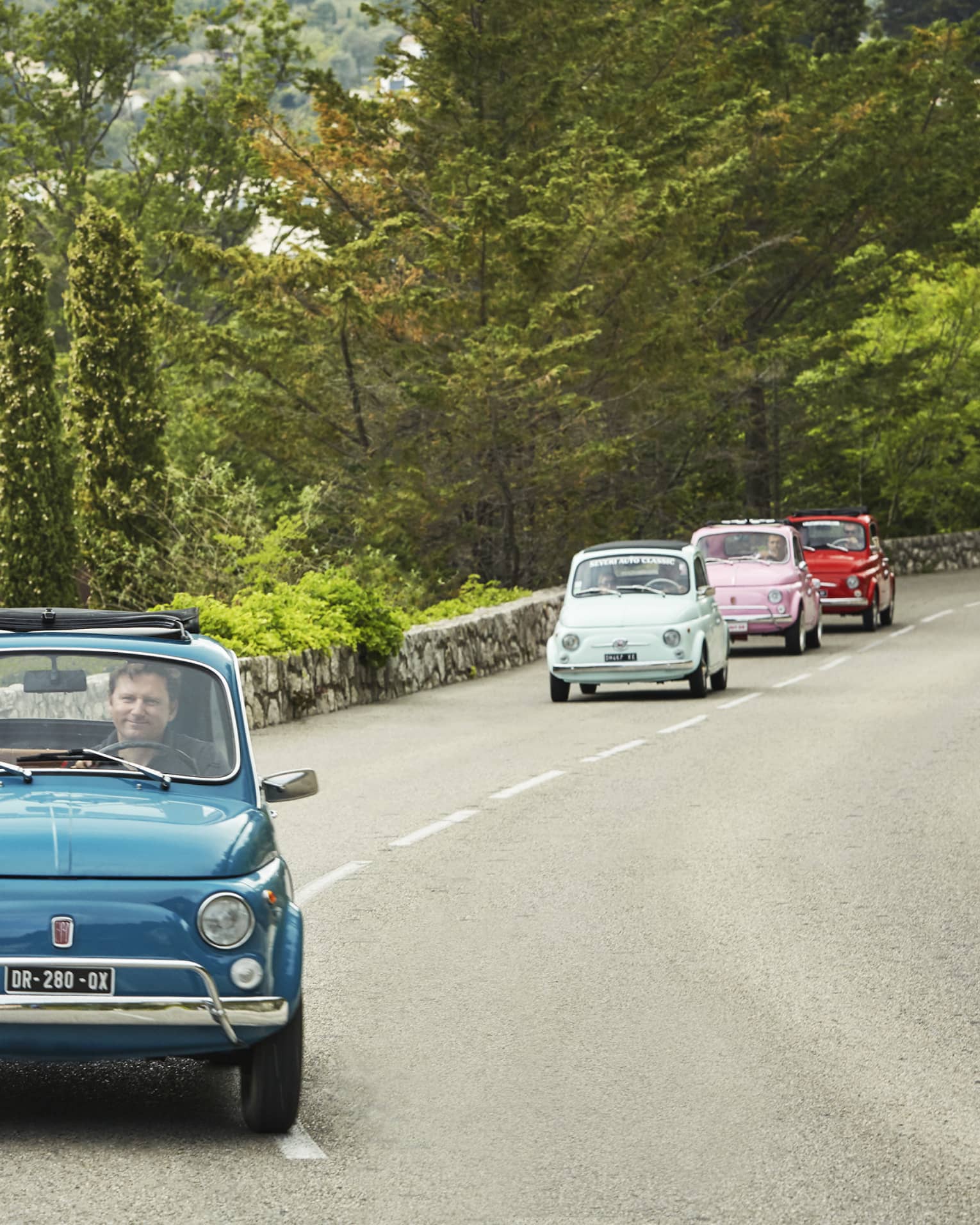Tourists drive colourful Fiat 500s down a road lined with a stone wall and greenery. The passenger of the first car waves.