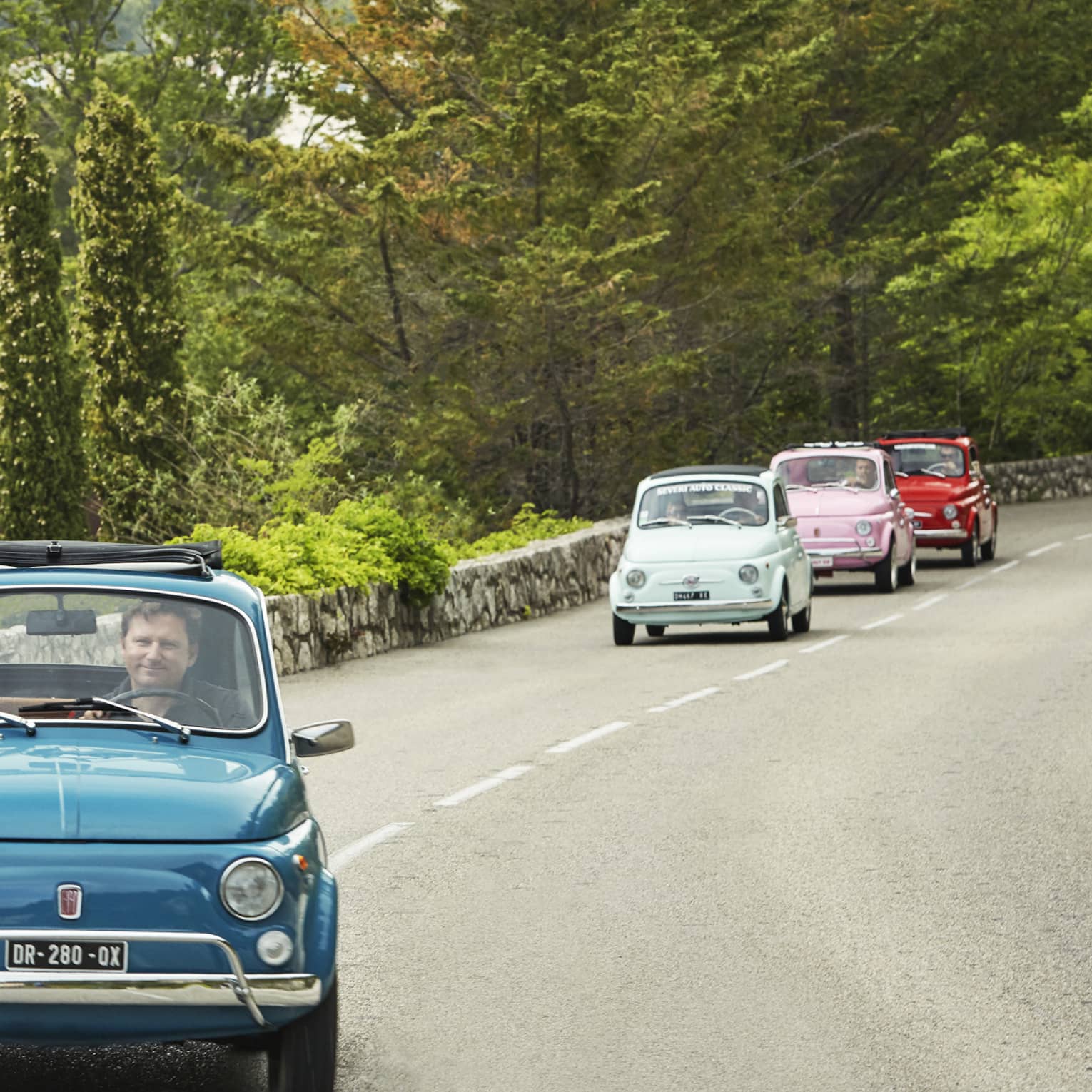 Tourists drive colourful Fiat 500s down a road lined with a stone wall and greenery. The passenger of the first car waves.