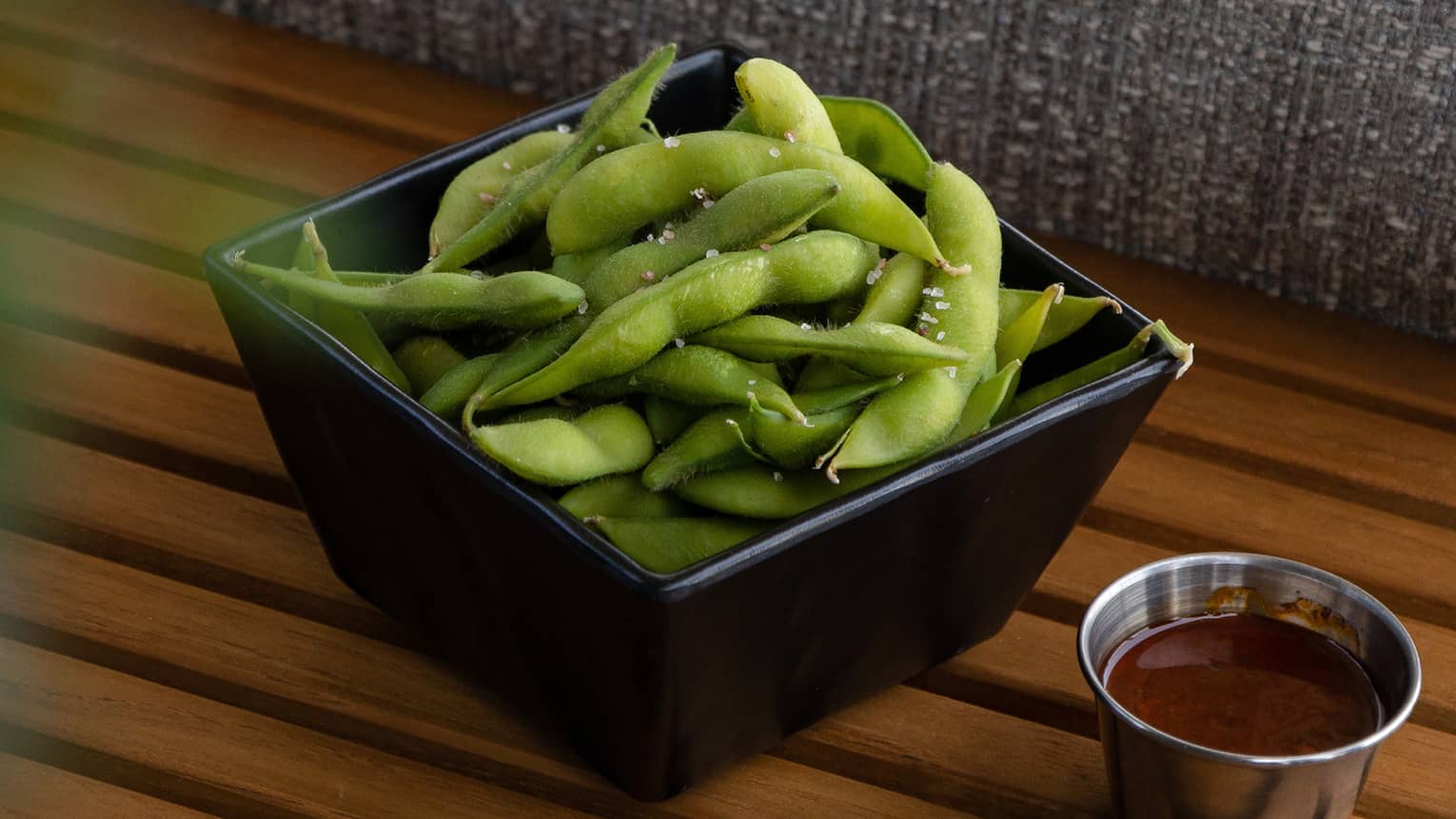 A black square bowl filled with steamed edamame beans sits next to a small silver dip bowl filled with sauce on a slated wood bench
