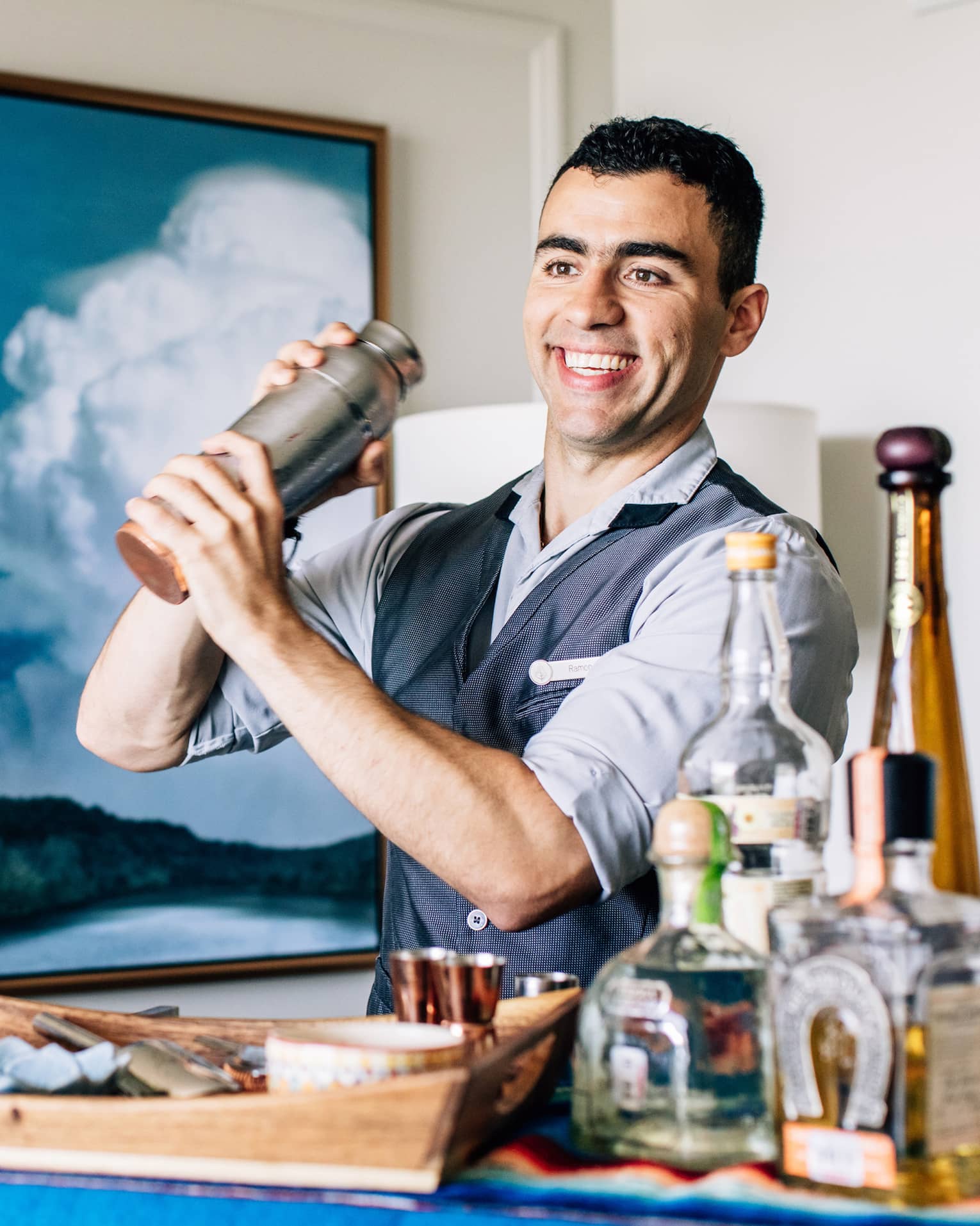 A bartender shaking a cocktail with liquor bottles on a cart in front of him.