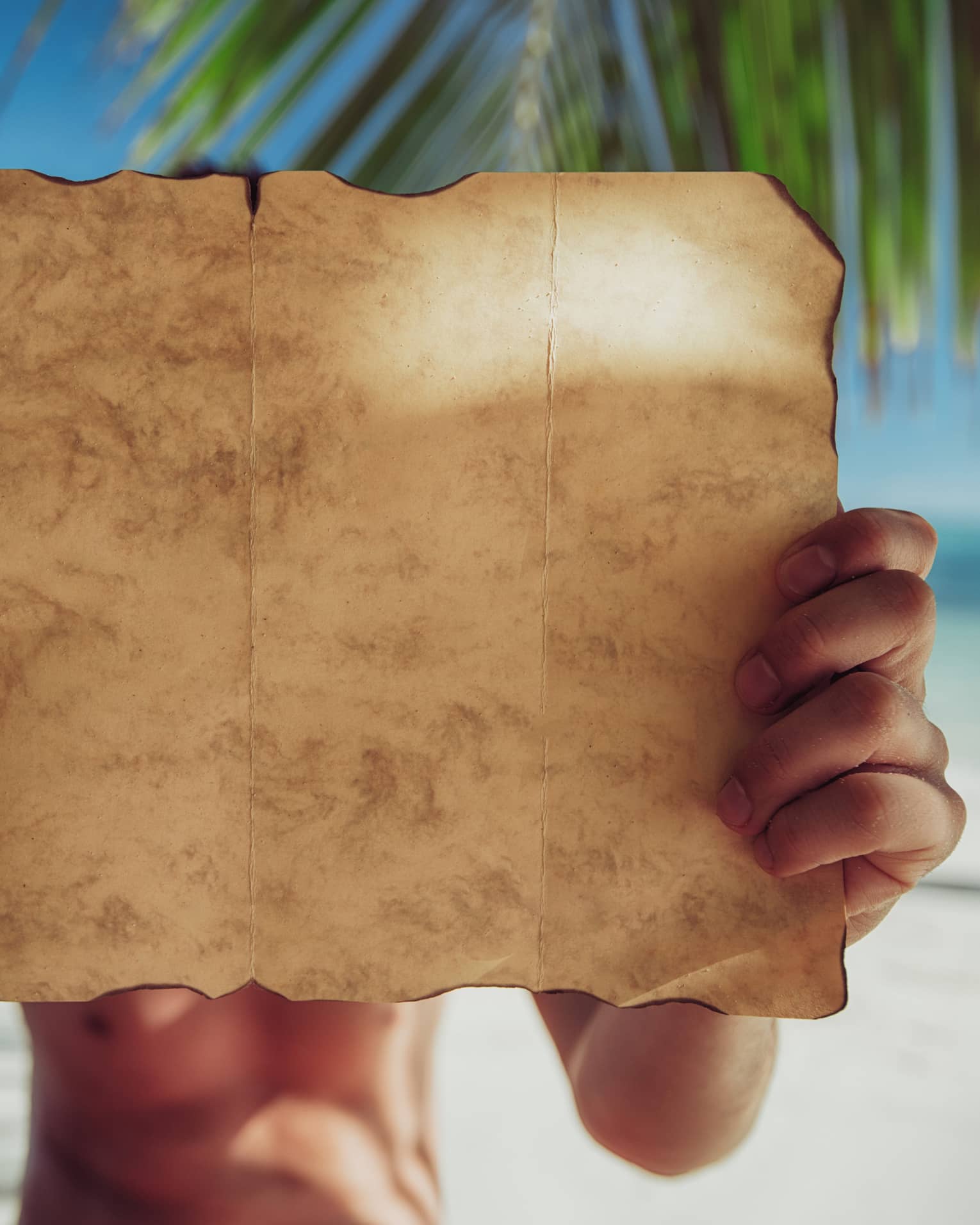 A close-up of a piece of parchment paper, yellow with age and burnt around the edges, being examined on a sandy beach.