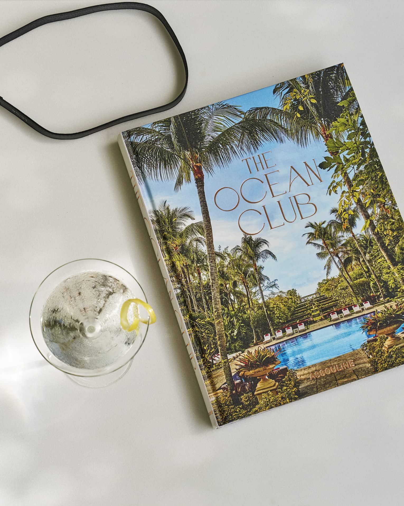 The Ocean Club Assouline book on a white surface next to a martini glass with a lemon twist garnish and camera